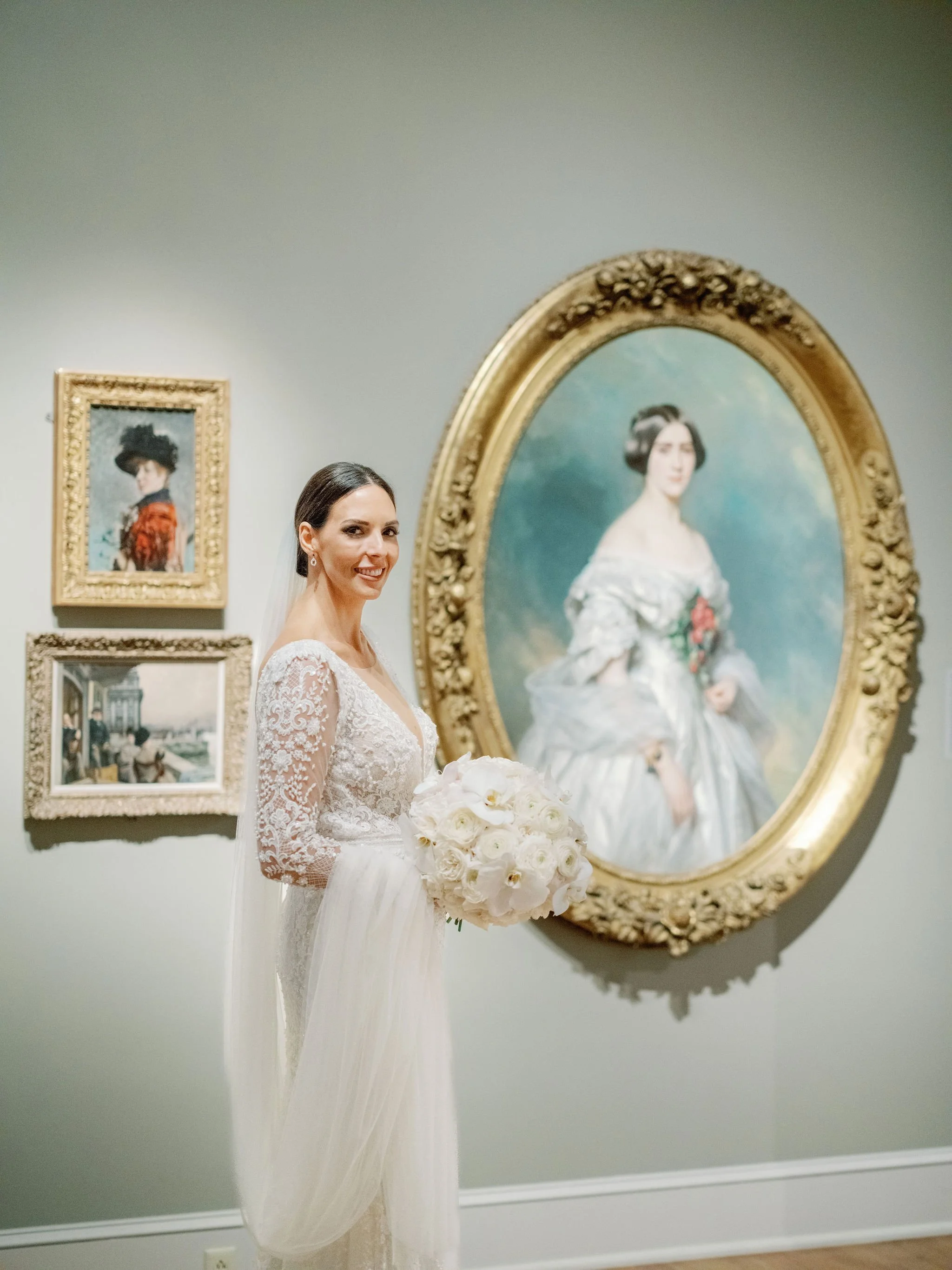 A bride in a lace wedding dress holding a bouquet of white roses, standing beside framed artwork and a large oval portrait of a woman in a white dress with dark hair, inside The New Orleans Museum of Art.