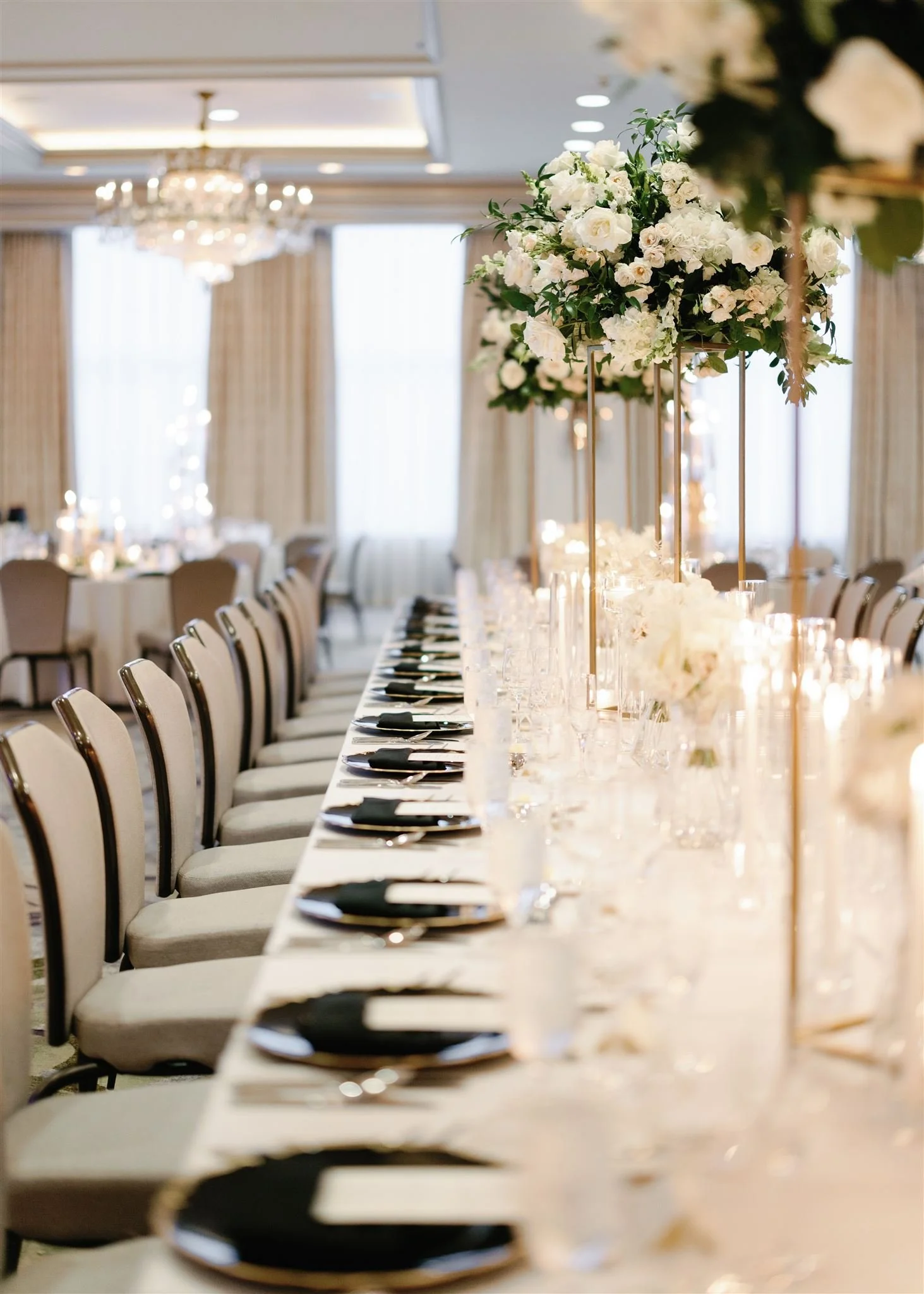 Elegant banquet hall with a long table set with black napkins, glassware, and floral centerpieces, with chandeliers and large windows with curtains in the background.