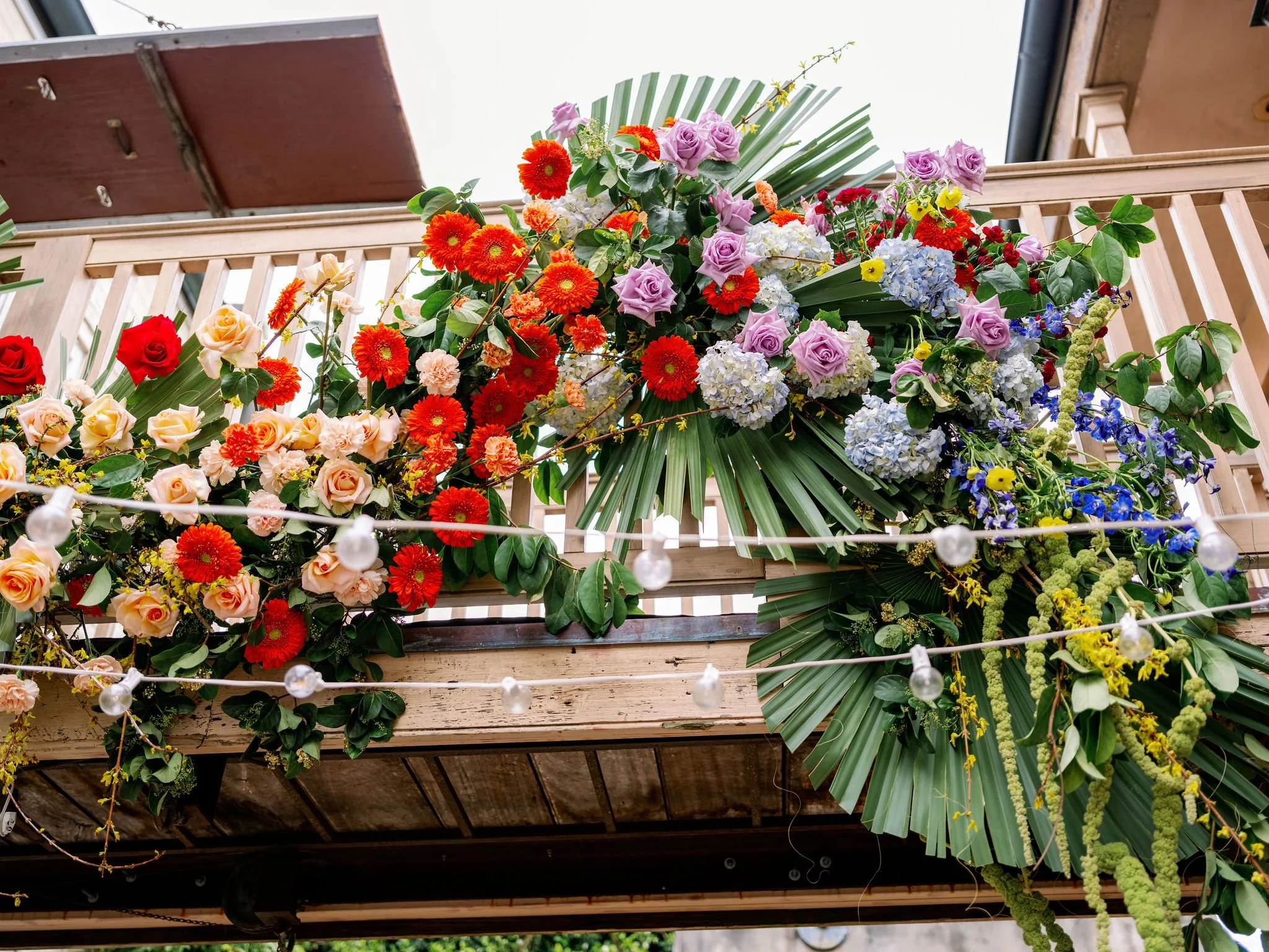Colorful ceremony florals with pink roses, orange and red gerbera daisies, white hydrangeas, blue delphiniums, and green foliage, displayed on a bride in the courtyard of Race and Religious. Designed by the best wedding planner in New Orleans