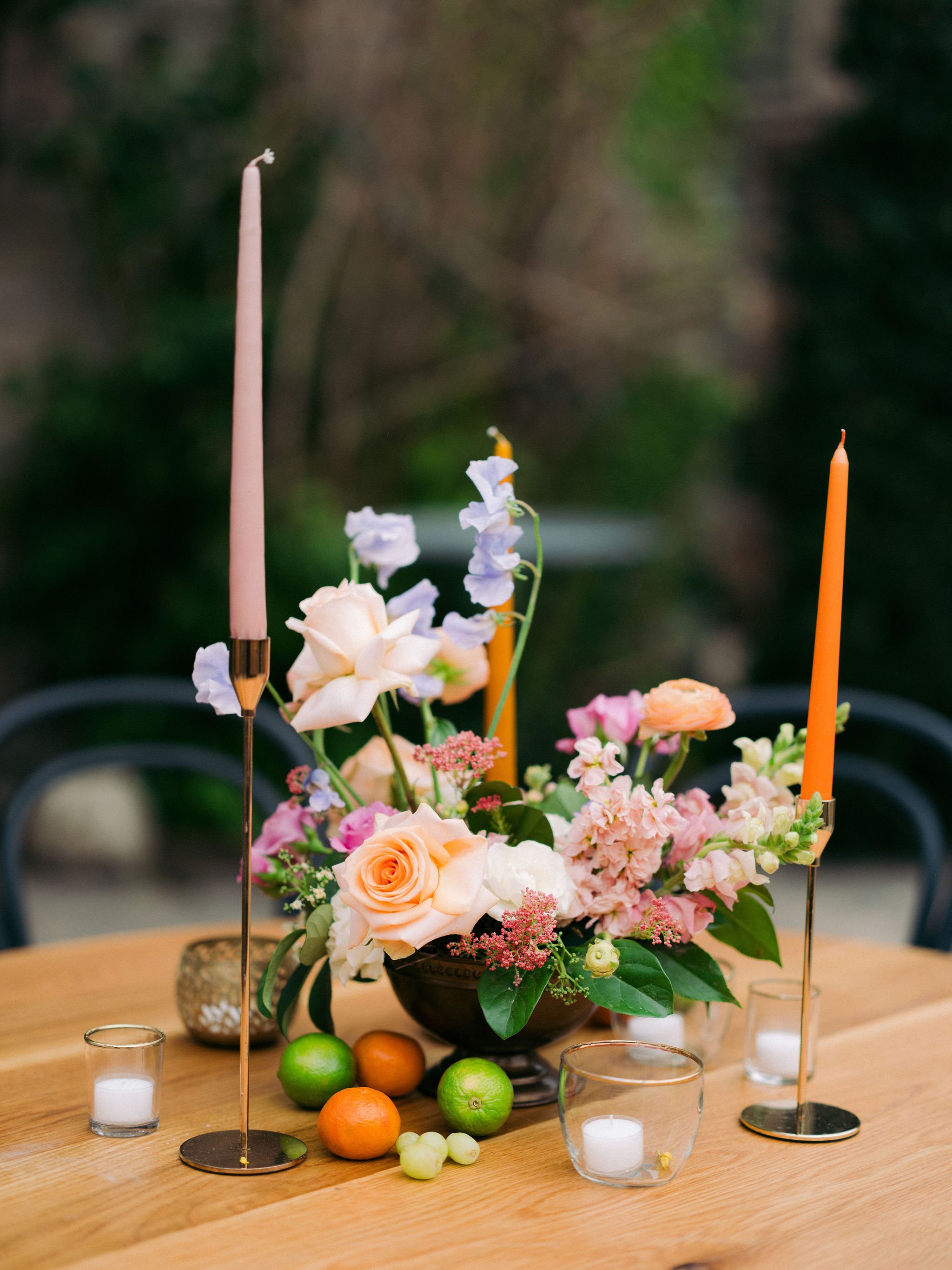 A floral arrangement with candles, candles, and fruits on a wooden table.