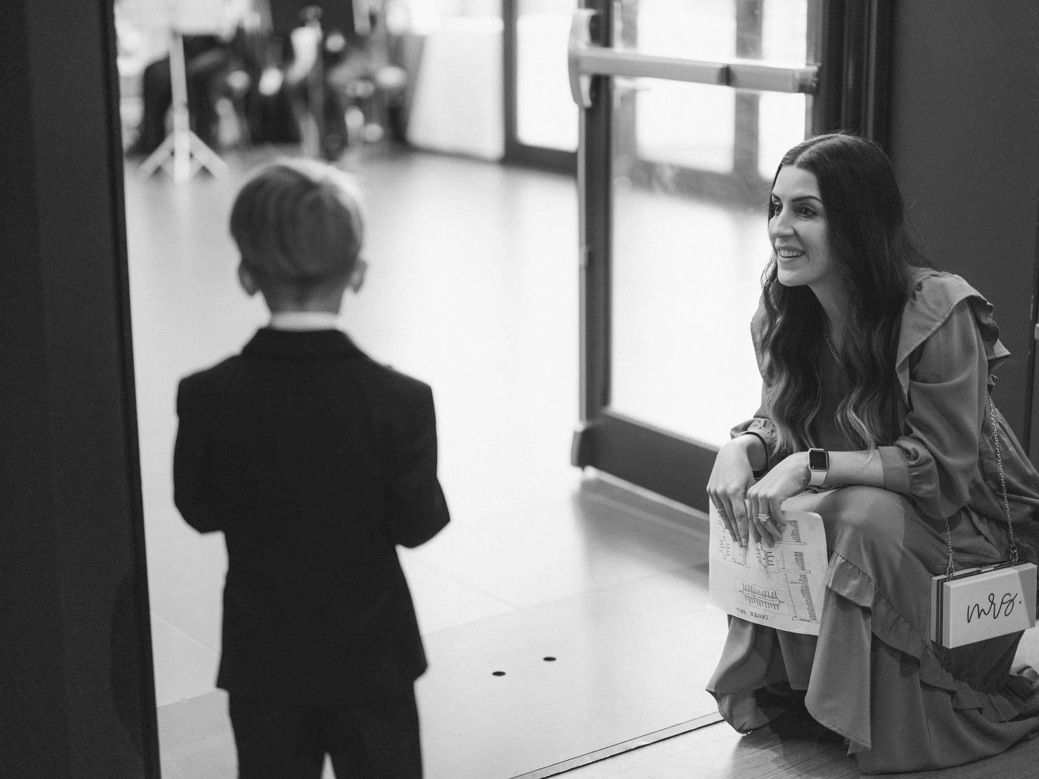 A woman with long dark hair smiling and leaning on a railing, holding papers and a clipboard, as she talks to a young boy dressed in a dark suit with short hair, inside a building near a glass door.