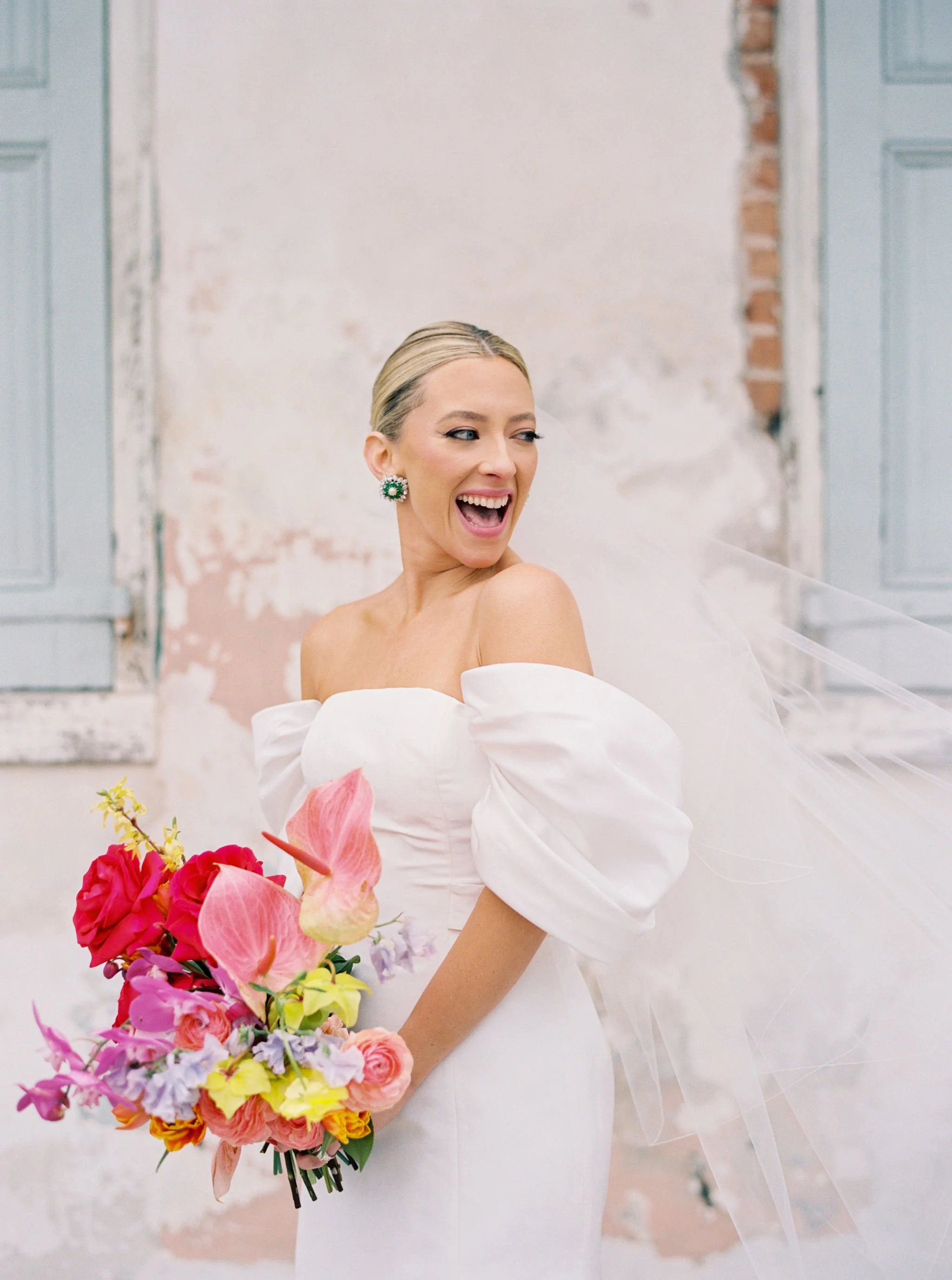 A chic bride in a white off-the-shoulder dress holding a colorful bouquet of flowers, standing in front of a textured white wall with blue doors at Race and Religious in New Orleans.