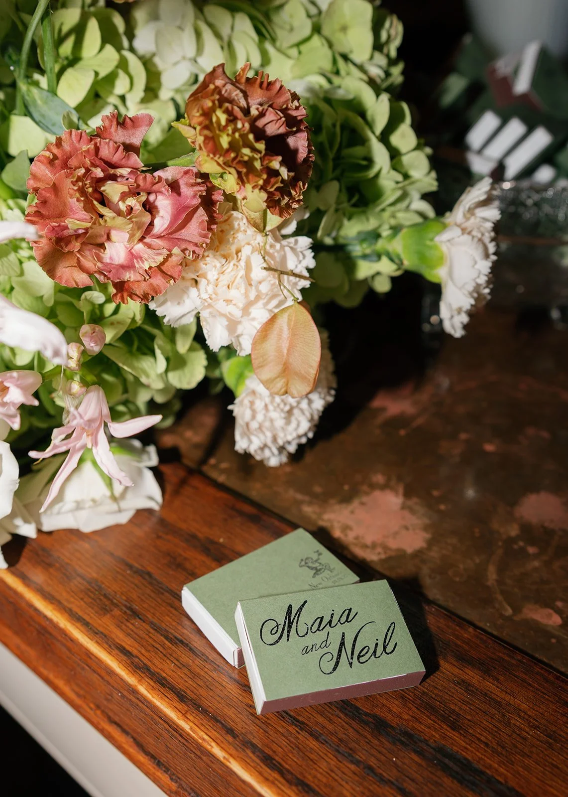 A floral arrangement with pink, brown, and white flowers on a wooden table. There is a small green card with black and cursive writing that says 'Maia and Neil'.