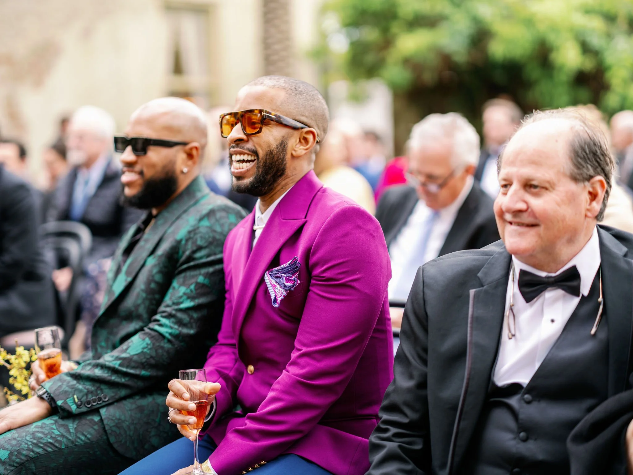 People dressed in formal attire, including a man in a purple suit and others in tuxedos, sitting at an outdoor event, smiling and holding glasses of champagne.