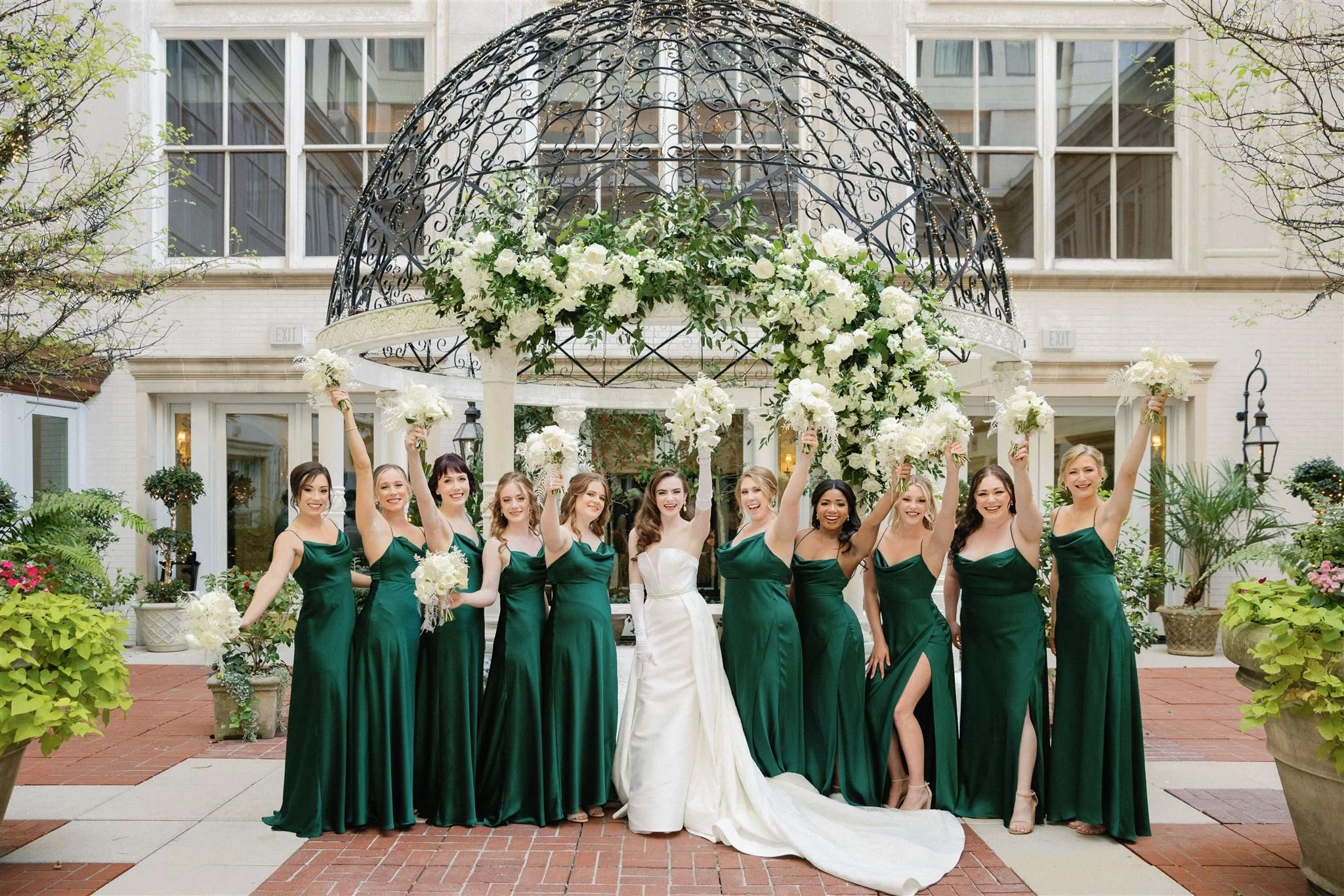A bride in a white wedding dress and a group of women in matching emerald green dresses celebrating at a wedding reception outside a building adorned with a floral arch and lanterns.