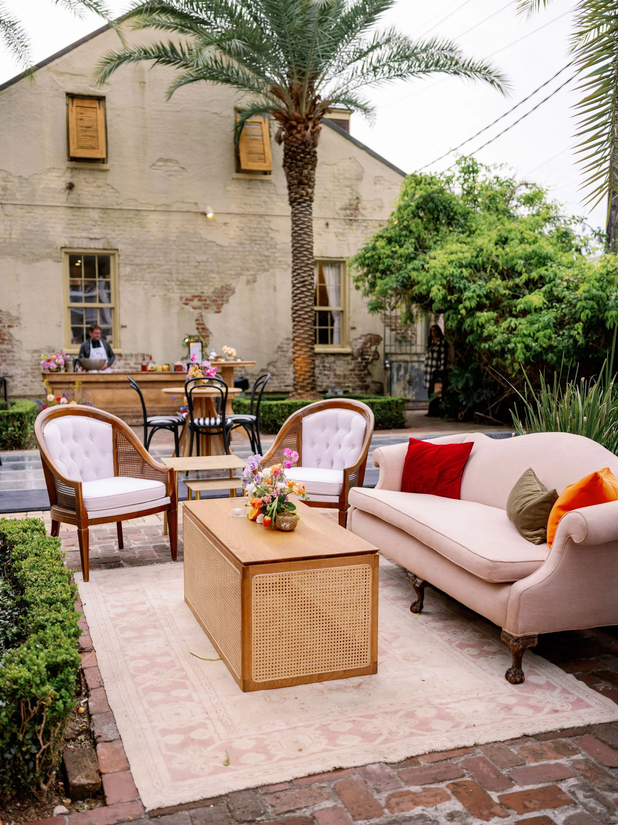 Outdoor patio with vintage furniture, including a pink sofa with colorful pillows, a wooden coffee table with a flower arrangement, and black and white chairs. The space features a pinkish rug on a brick patio, lush greenery, and a large palm tree. I
