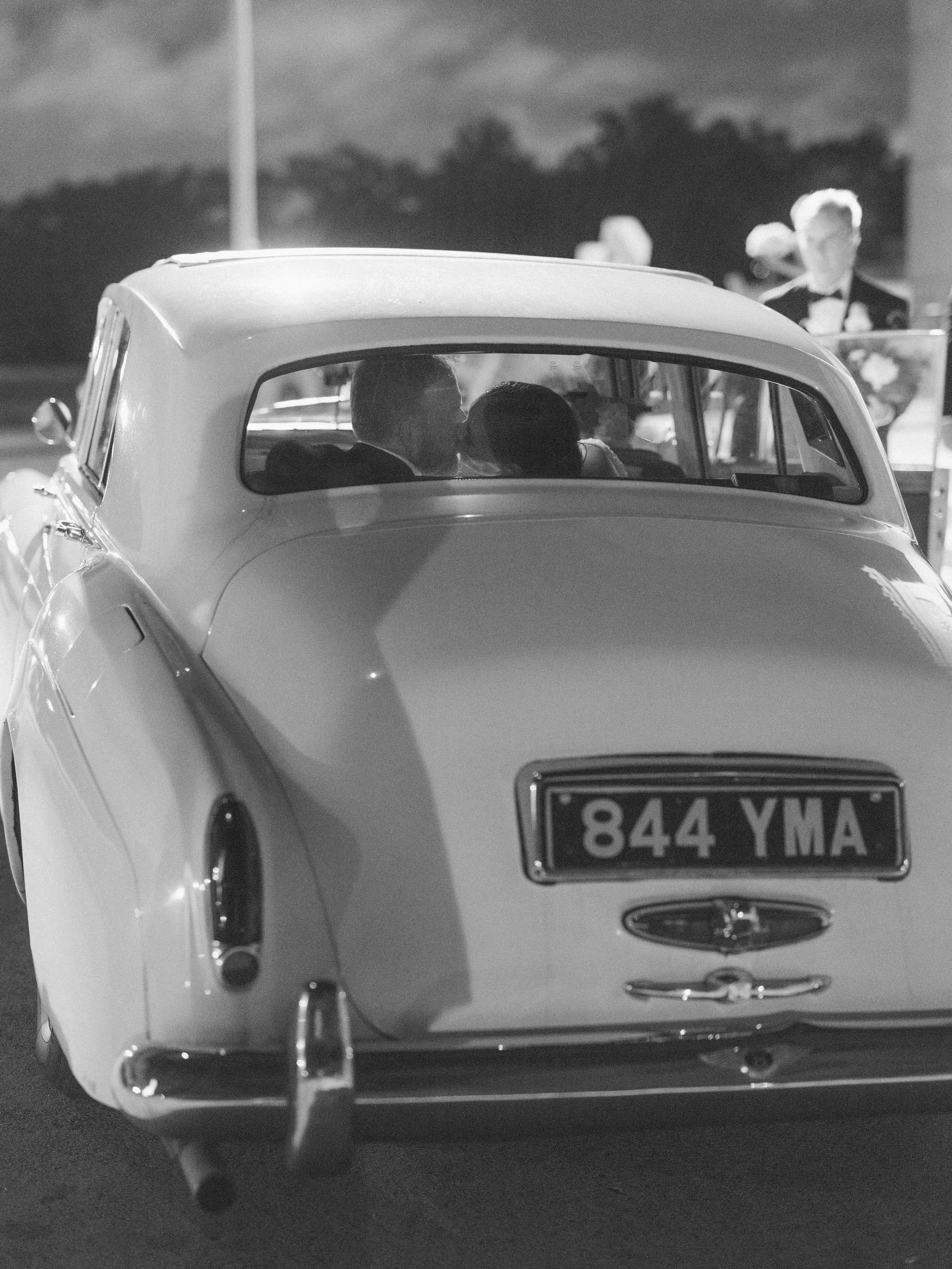 A vintage car with a partial view of the license plate 844 YMA, parked outdoors at night. The rear of the car is visible, with a person inside looking out the window. In the background, a man in a suit with a bowtie stands near a table with flowers, 