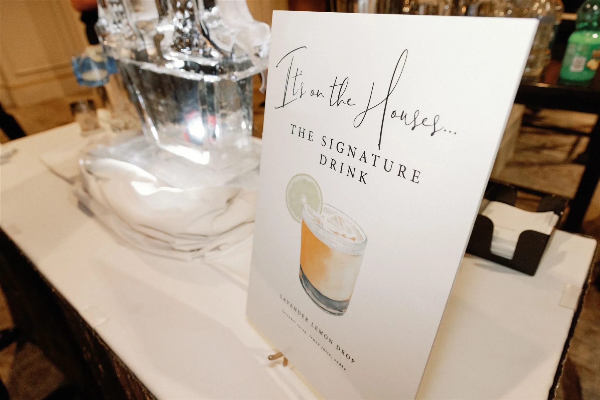 A sign on a table indicating 'It’s on the House... The Signatures Drink' with an illustration of an orange cocktail garnished with a lime slice, set on a white tablecloth with a large glass container filled with ice and a green bottle in the backgrou