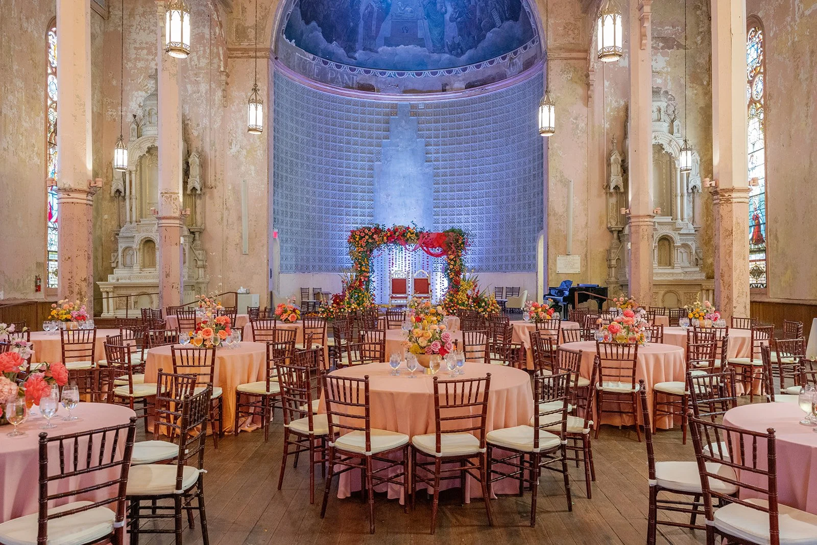 Inside a church decorated for a wedding with round tables covered in pink tablecloths, floral centerpieces, and chairs around each table. At the front, there is an arch decorated with flowers and a backdrop with two chairs for the bride and groom. Th