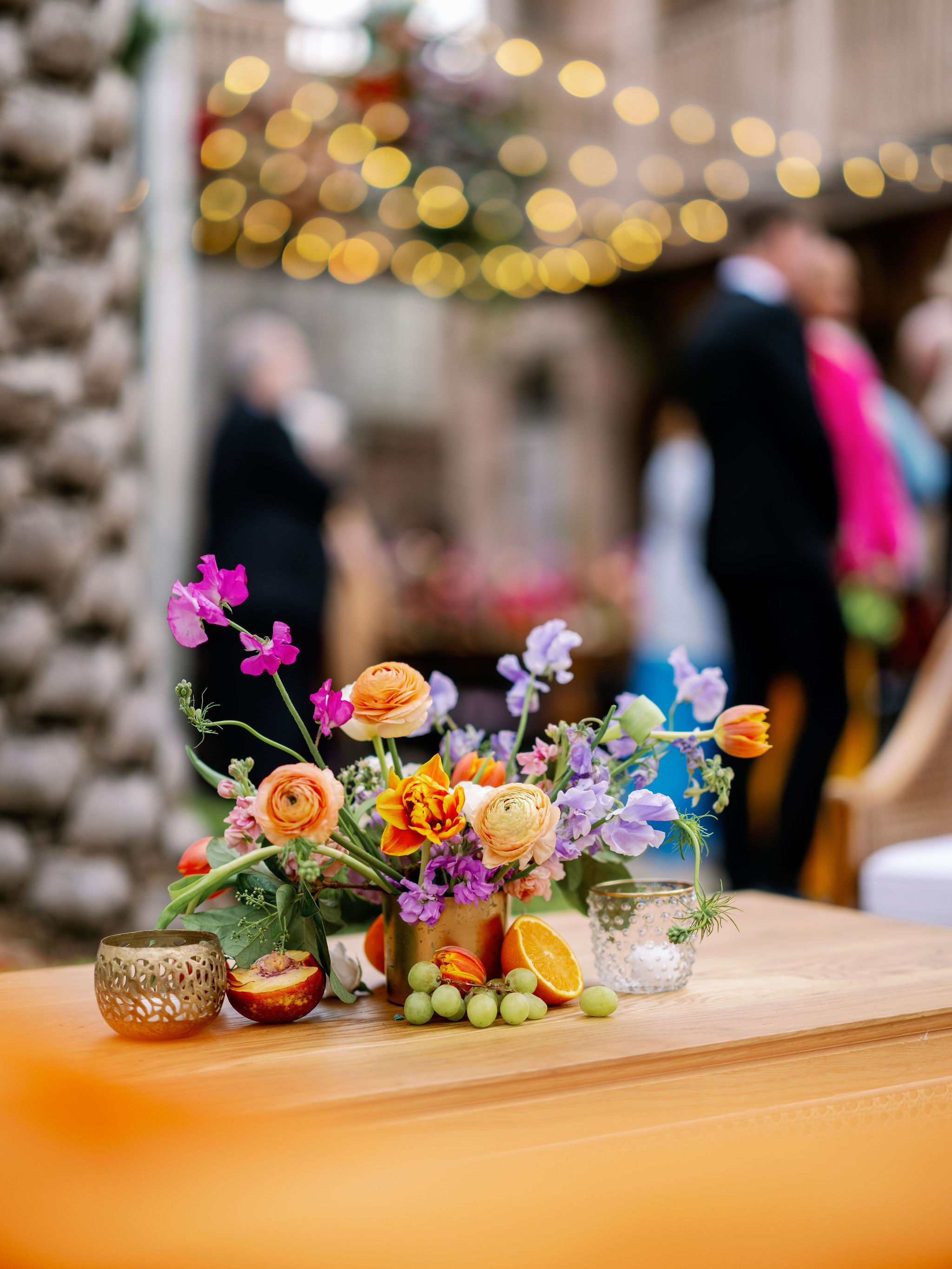 A colorful flower arrangement on a wooden table with oranges, grapes, and decorative candle holders in the foreground, blurred guests in the background at a festive gathering.