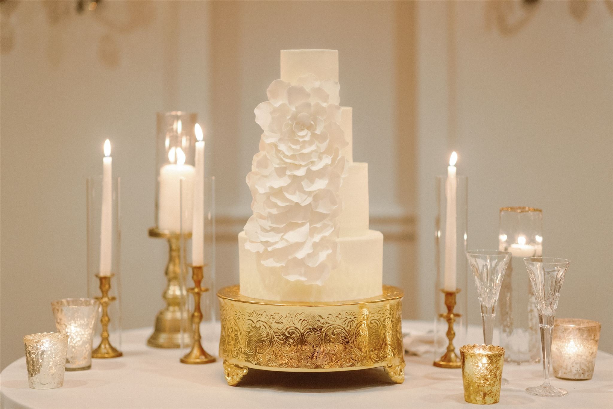 A multi-tiered white cake with large white flower decorations, displayed on a gold cake stand, surrounded by gold and clear candle holders with lit candles, on a white tablecloth.