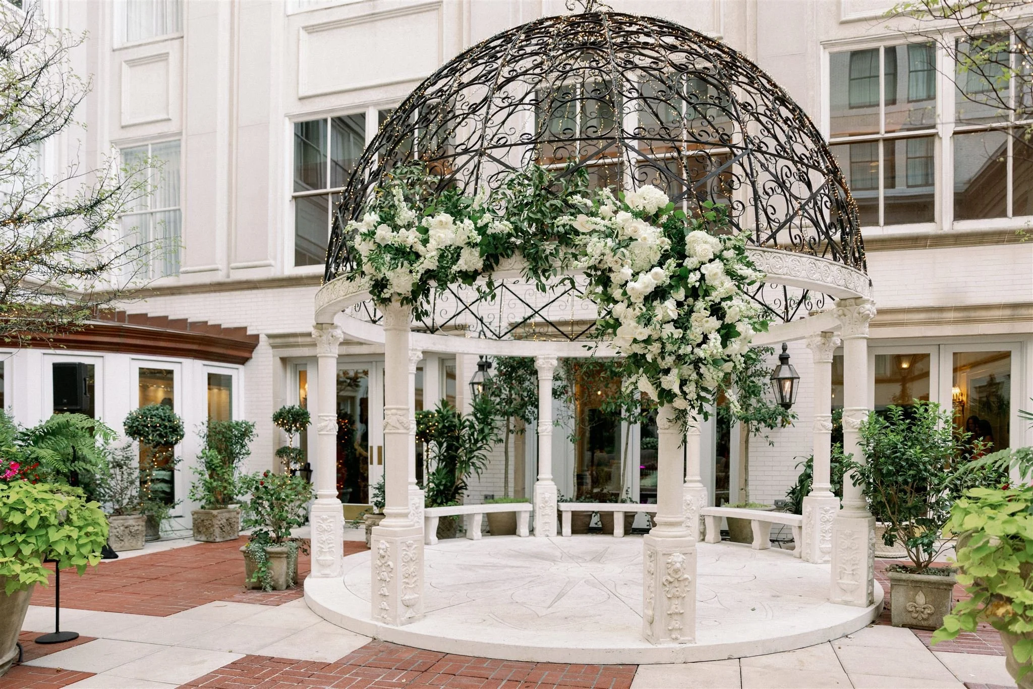A white circular wedding altar decorated with white flowers and greenery is set in an outdoor courtyard with a building in the background.