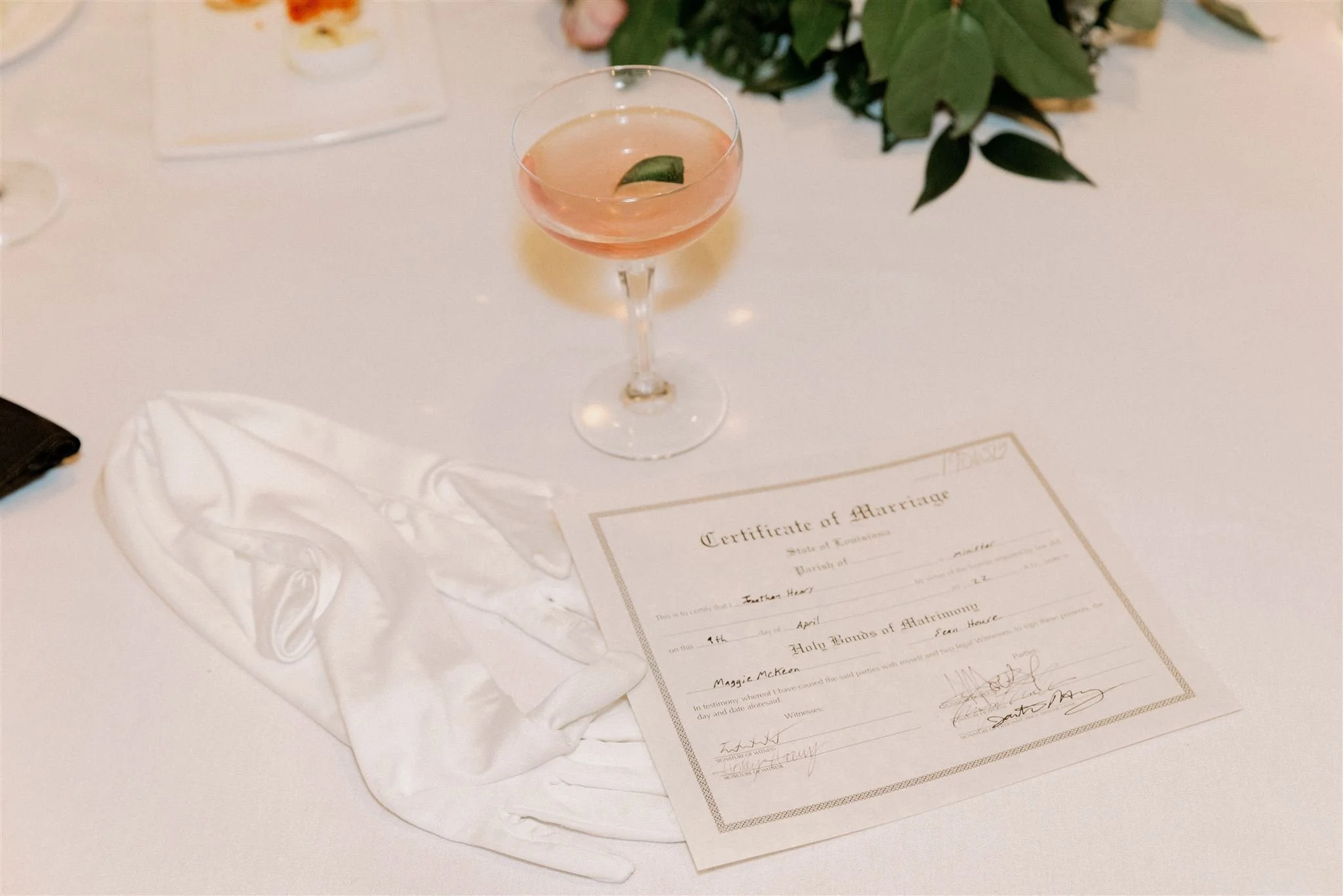 A wedding certificate and a glass of pink cocktail with a green leaf garnish on a white table, with a floral arrangement in the background.