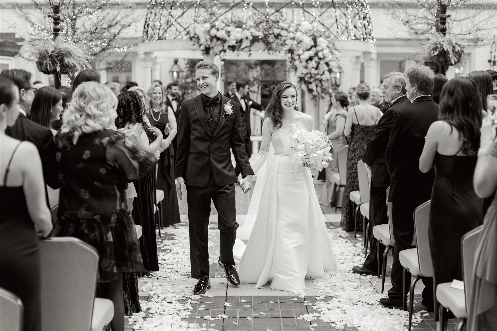 A bride and groom walking hand in hand down the aisle at their wedding ceremony, surrounded by seated guests and outdoor decorations.