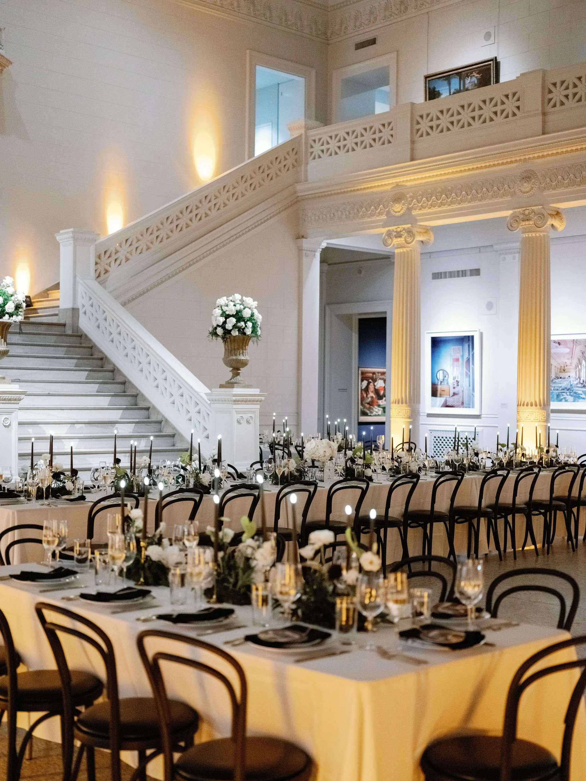 Elegant banquet hall with round tables set with white tablecloths, black chairs, floral centerpieces, and candlelit dinnerware, with a grand staircase, ornate columns, and artwork on the walls.