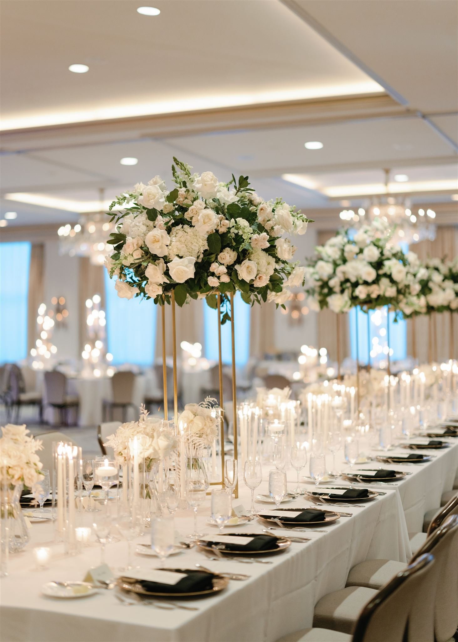 Elegant banquet table decorated with tall floral arrangements of white and blush roses with greenery, set for a formal event in a brightly lit ballroom.