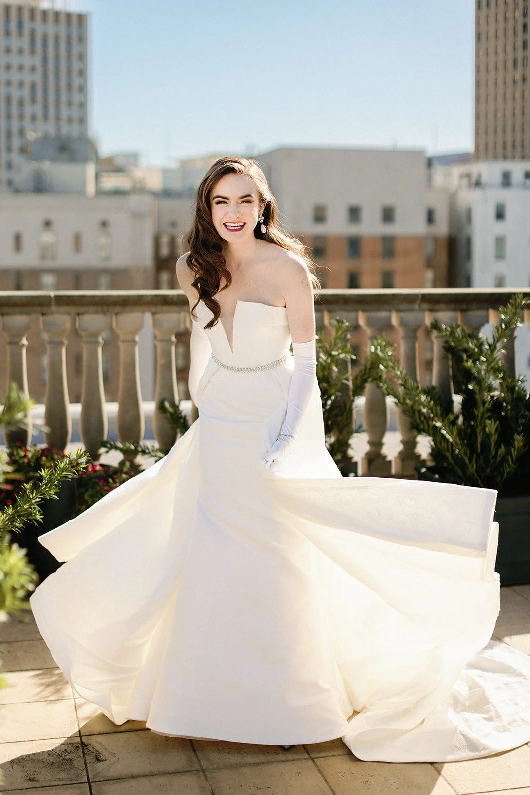 A woman in a white wedding dress standing outdoors on a balcony, smiling and winking. The background shows tall buildings and a clear sky.
