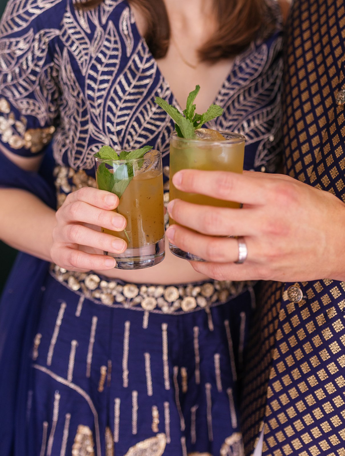 Two people holding cocktails garnished with mint leaves, one wearing traditional Indian attire with intricate embroidery, the other in a patterned outfit.