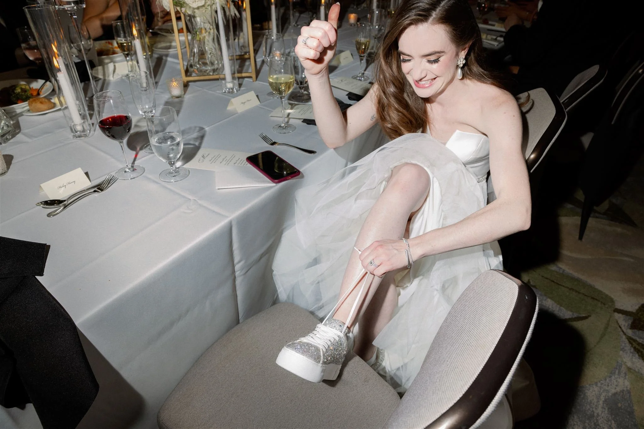 A woman in a white dress with sparkly shoes sitting at a dinner table, smiling and making a gesture with her hand. The table is set with wine glasses, water glasses, plates, silverware, menus, and candles.