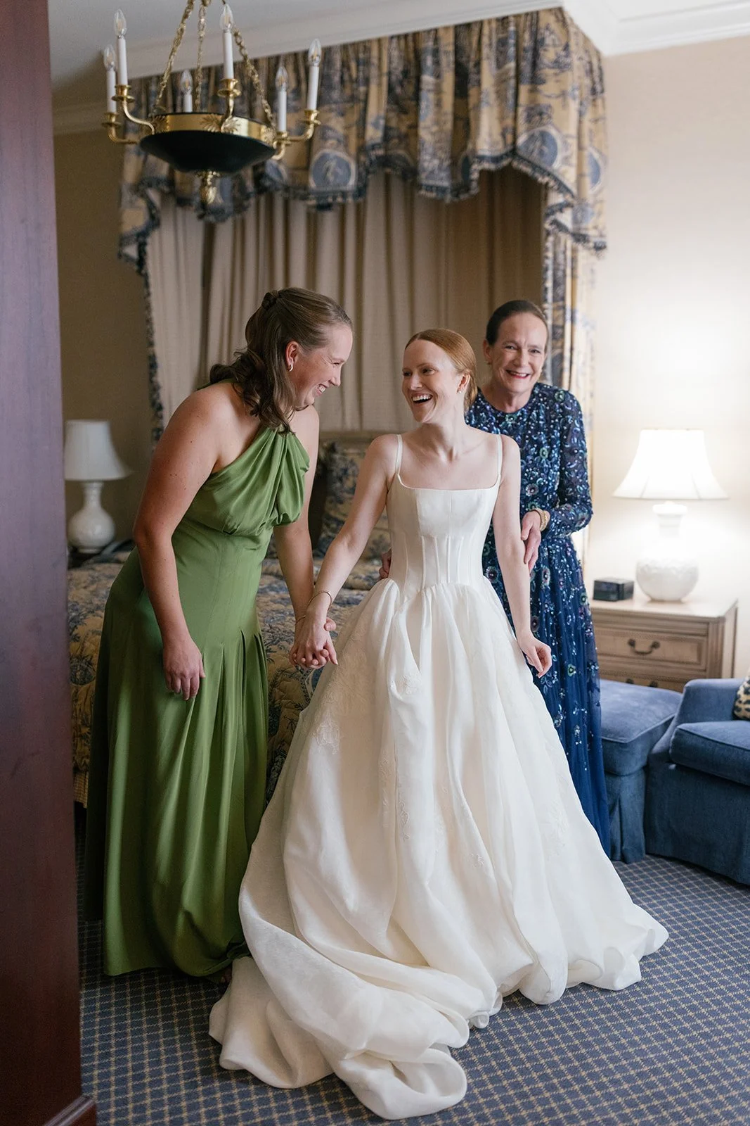 A bride in a white wedding dress is holding hands with two women, one in a green dress and the other in a blue dress, as they smile and laugh in a warmly lit room with elegant curtains and a chandelier.