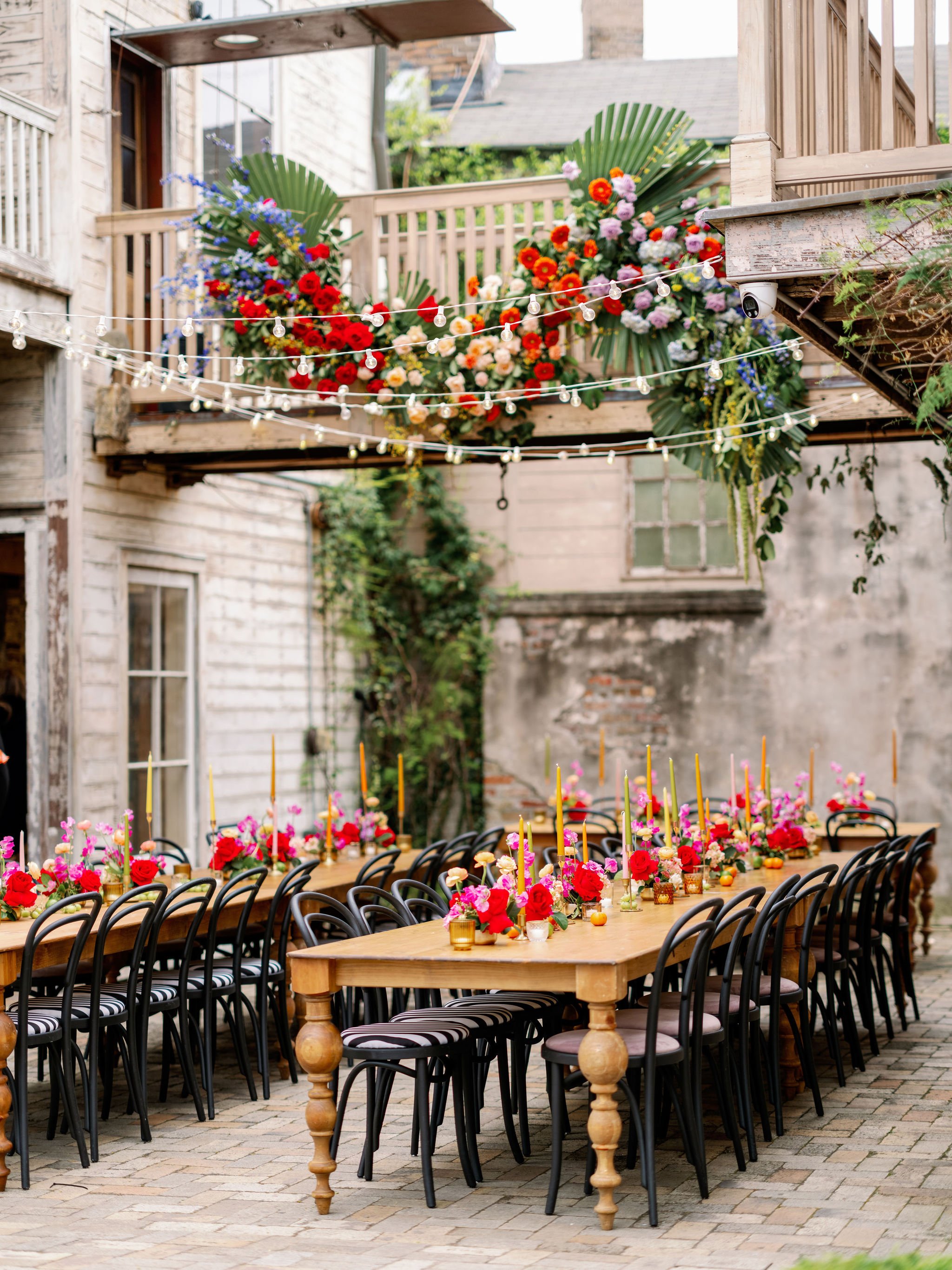 A long wooden table set for an outdoor celebration with colorful flowers, candles, and gold accents, surrounded by black chairs with striped cushions. String lights hang above, and a wooden balcony with floral arrangements is in the background.