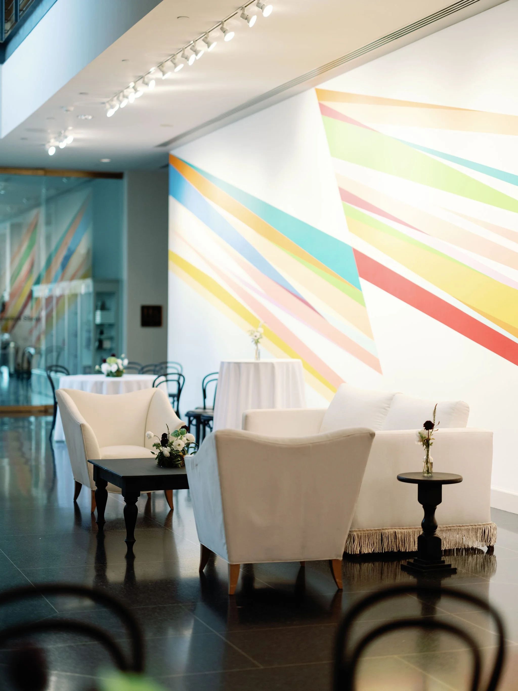 Elegant interior of a lounge or restaurant area with cream-colored armchairs, small black side tables with vases of flowers, and a colorful geometric mural on the wall. In the background, there are round tables with white tablecloths and floral cente