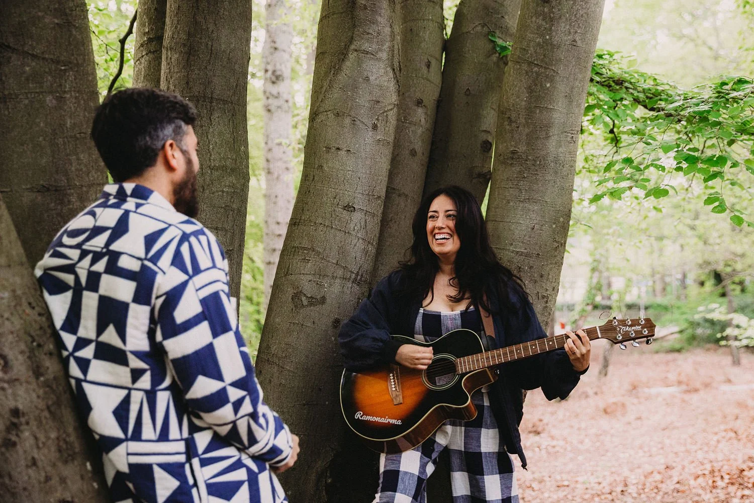 A woman playing guitar and singing outdoors while two men listen near trees in a forest park.