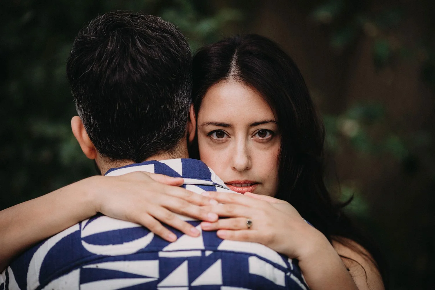 A woman with dark hair and expressive eyes hugging a man with dark hair from behind, outdoors with a blurred natural background.