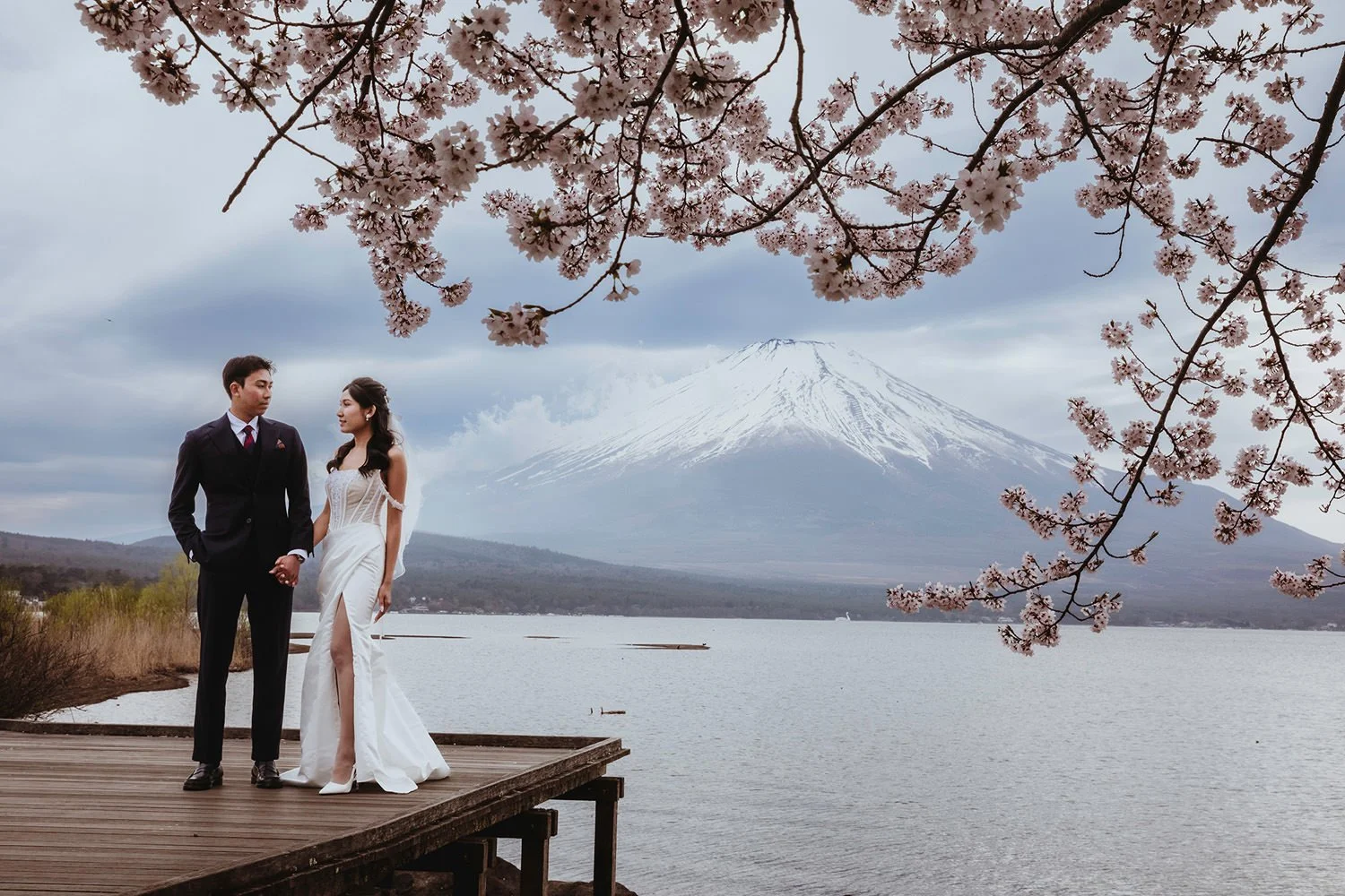 A couple in wedding attire holding hands on a wooden dock by a lake, with Mount Fuji and cherry blossoms in the background.