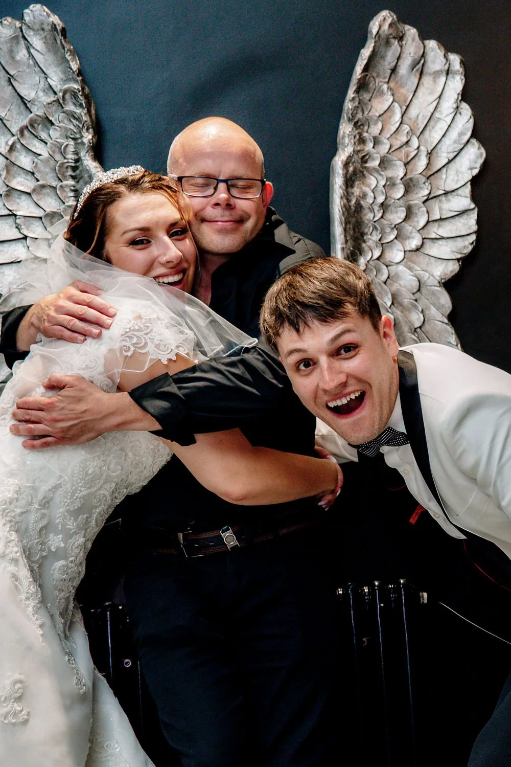 People hugging and smiling at a wedding reception, with angel wing decorations in the background.