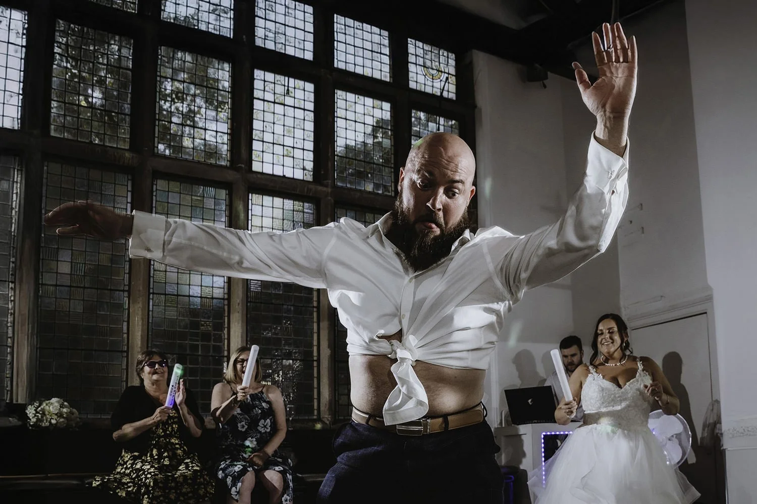A man with a beard and bald head wearing a white shirt tied at the stomach, dancing at a celebration. In the background, a woman in a wedding dress and other guests are smiling and holding glow sticks in a room with large stained glass windows.
