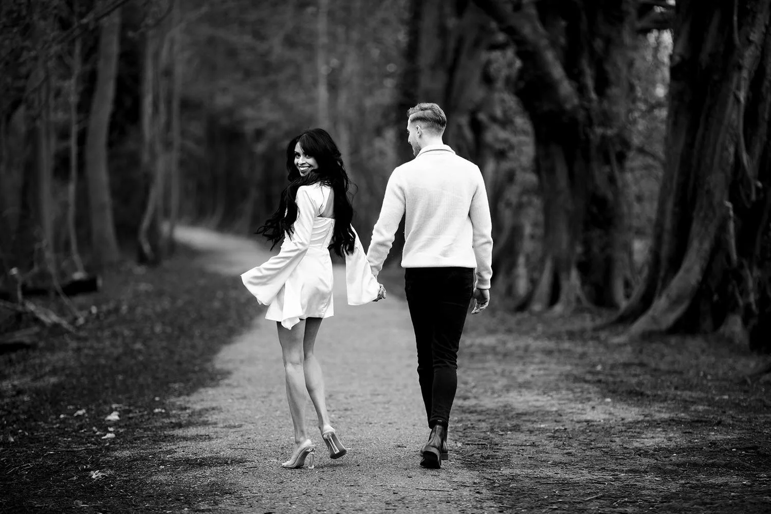 A black-and-white photo of engaged couple holding hands while walking on a dirt path through a wooded area, with the woman turning and smiling at the camera.