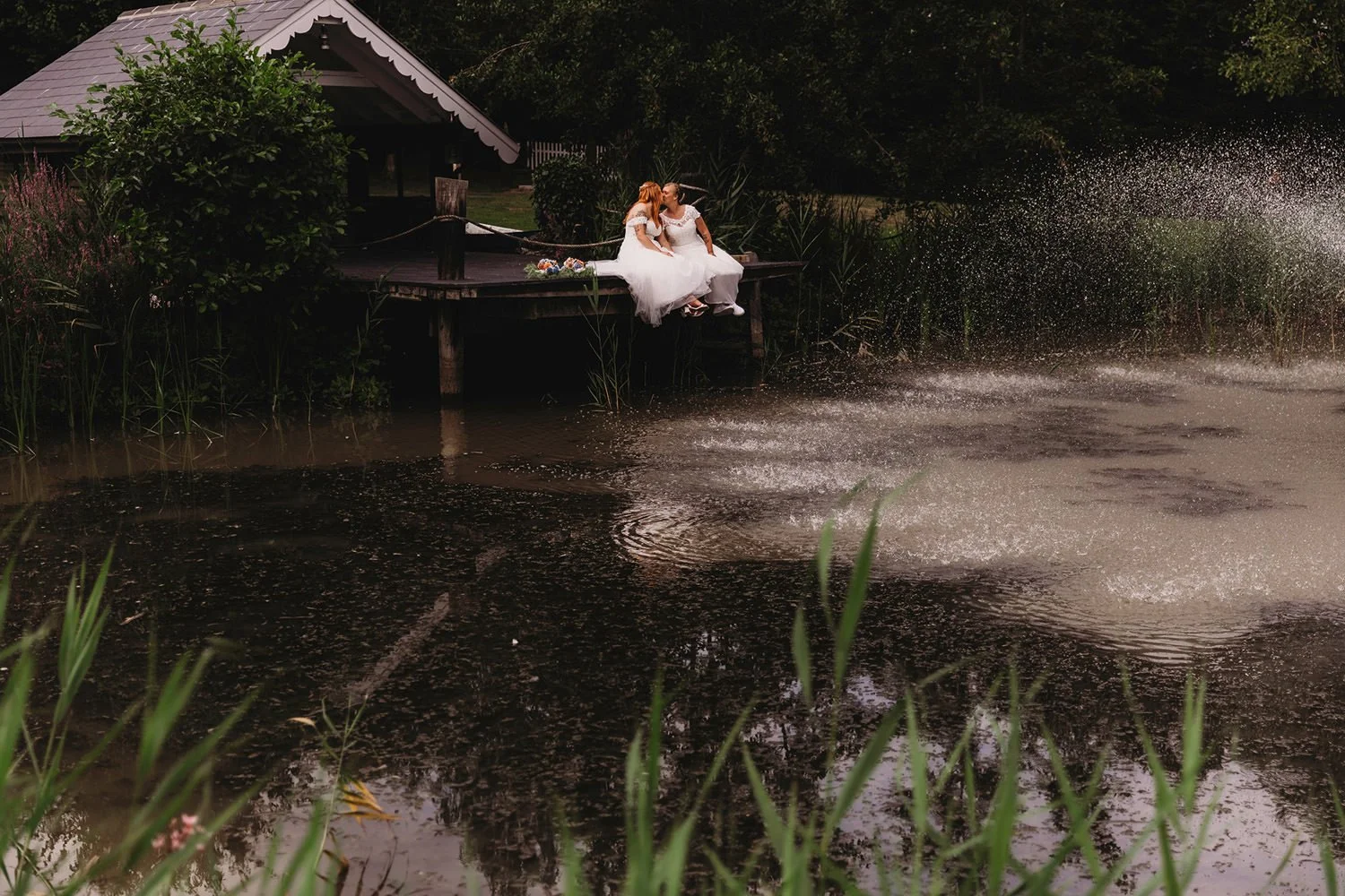 Two women in wedding dresses sitting on a wooden dock by a pond, sharing a kiss, with a small shed and greenery in the background, and water splashing in the pond.