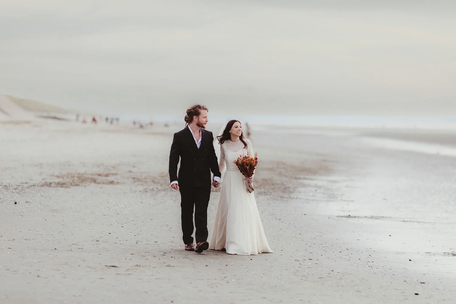 A newlywed couple walking hand in hand on a beach, with the bride holding a bouquet of flowers and smiling, the groom dressed in a black suit, with a blurred background of other people on the beach.