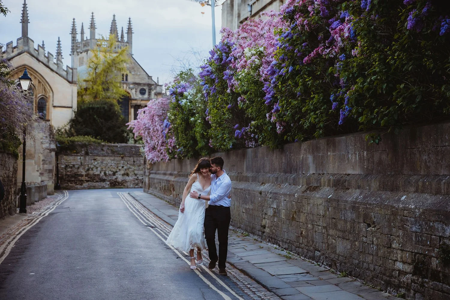 A couple walks on a narrow street lined with colorful flowers, with a historic building in the background.