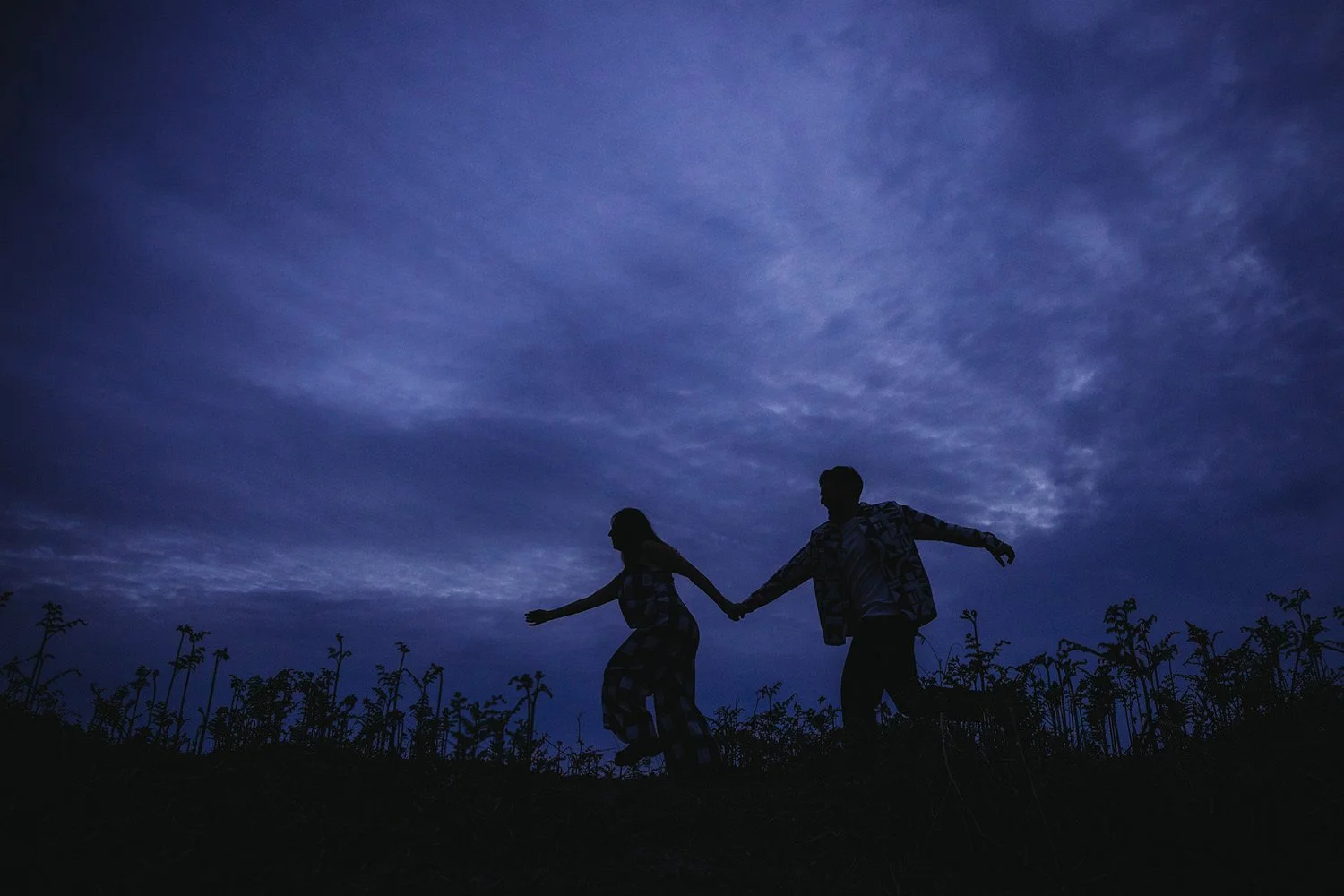 Silhouettes of a man and woman holding hands and playing on a hilltop at dusk with a deep blue sky and scattered clouds in the background.