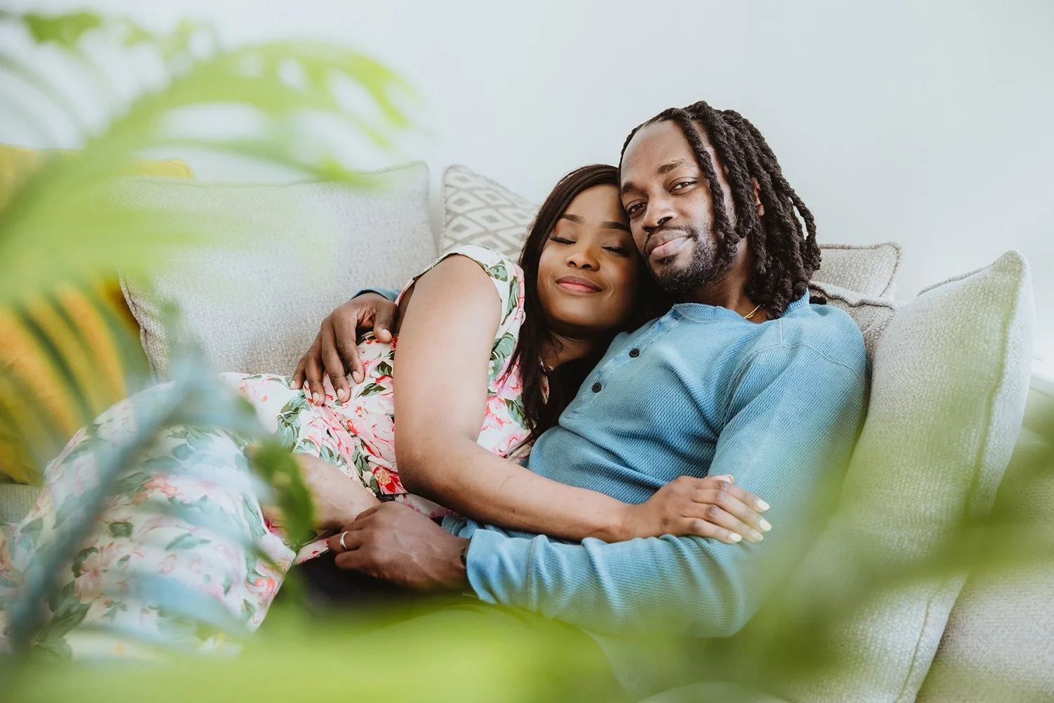 A couple sitting on a beige couch, embracing lovingly with closed eyes, surrounded by throws and cushions, indicating a cozy indoor setting.