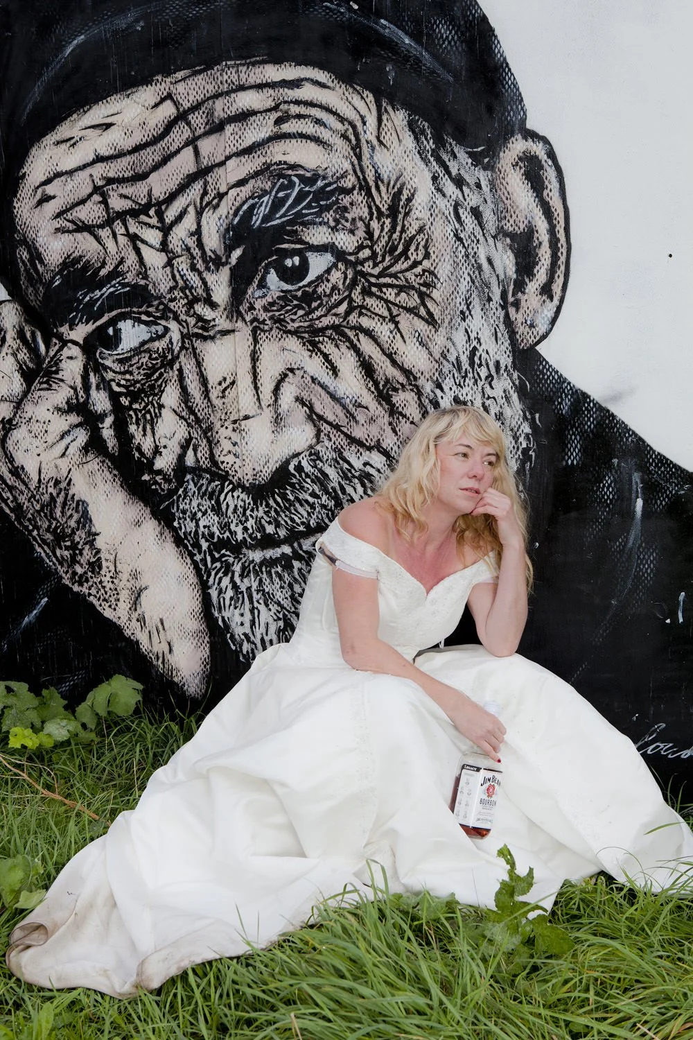 A woman in a white dress sitting on grass, holding a bottle of Jim Beam bourbon, with a large mural of an elderly man's face in the background.