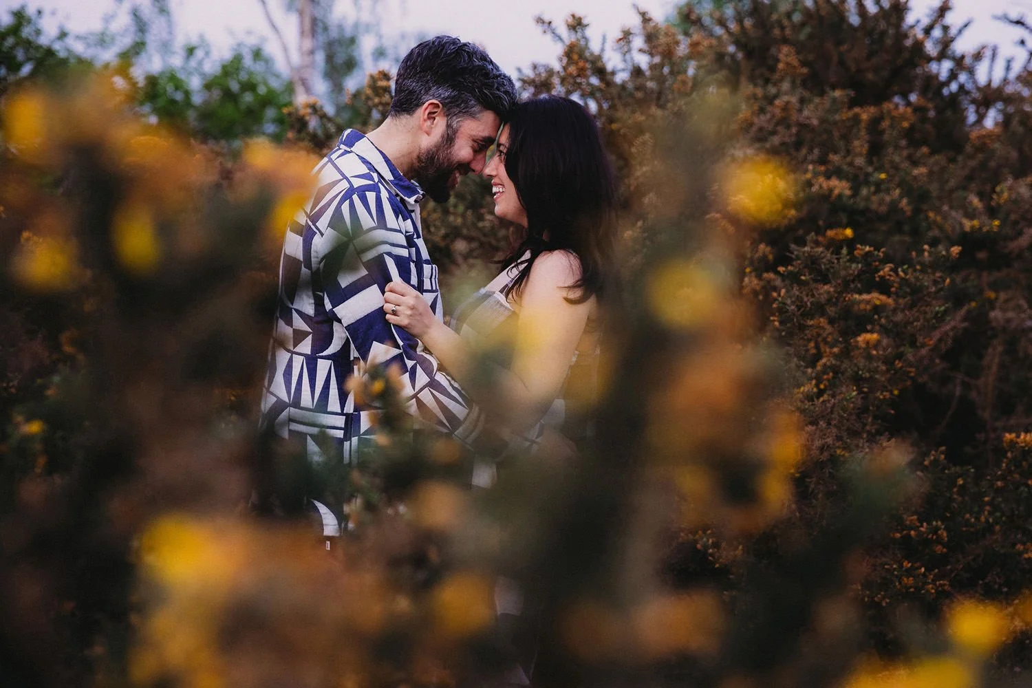 A man and woman standing close together outdoors during sunset, smiling and touching foreheads, surrounded by bushes with yellow flowers.