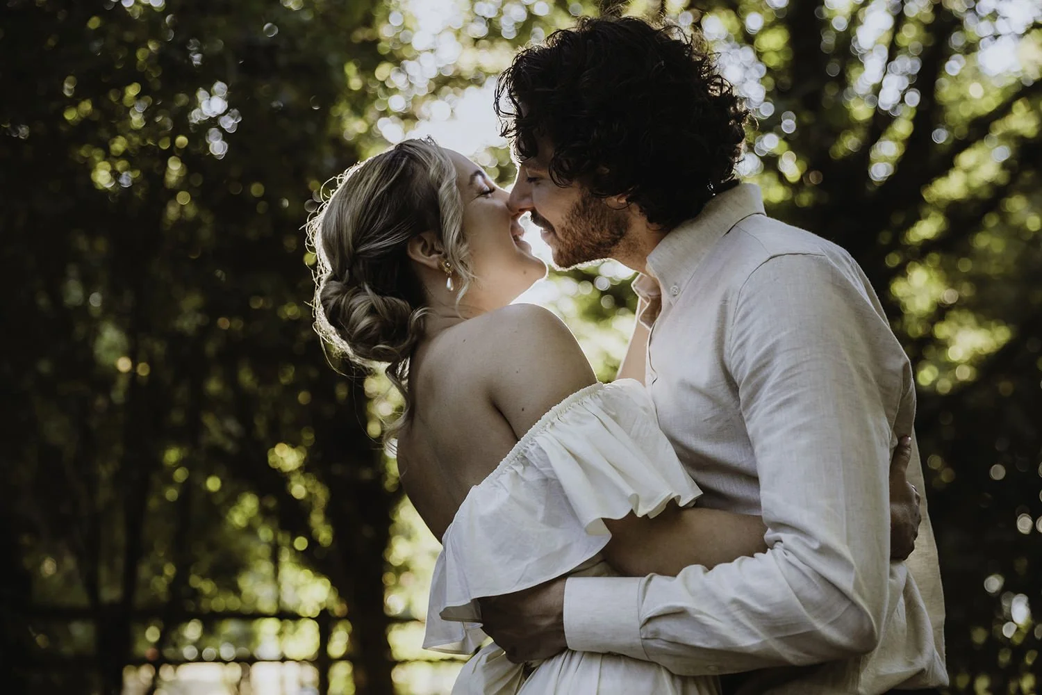 A couple standing closely together outdoors, about to kiss, with sunlight and trees in the background.