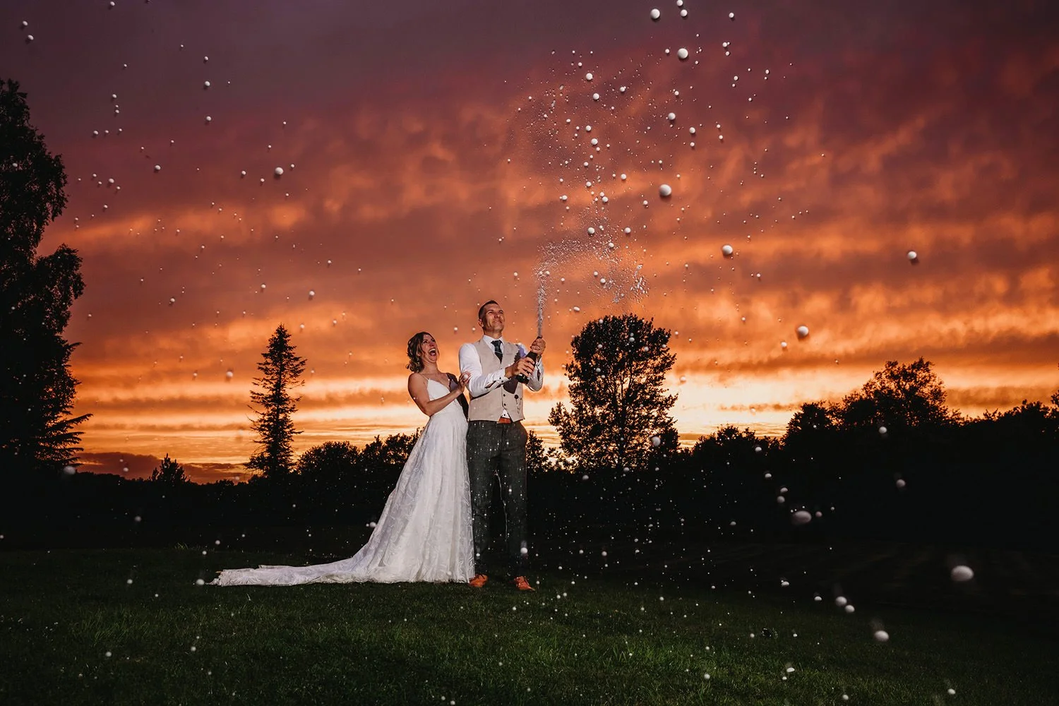 A newlywed couple celebrating with champagne outdoors at sunset, with tan and orange clouds in the sky and trees in the background.