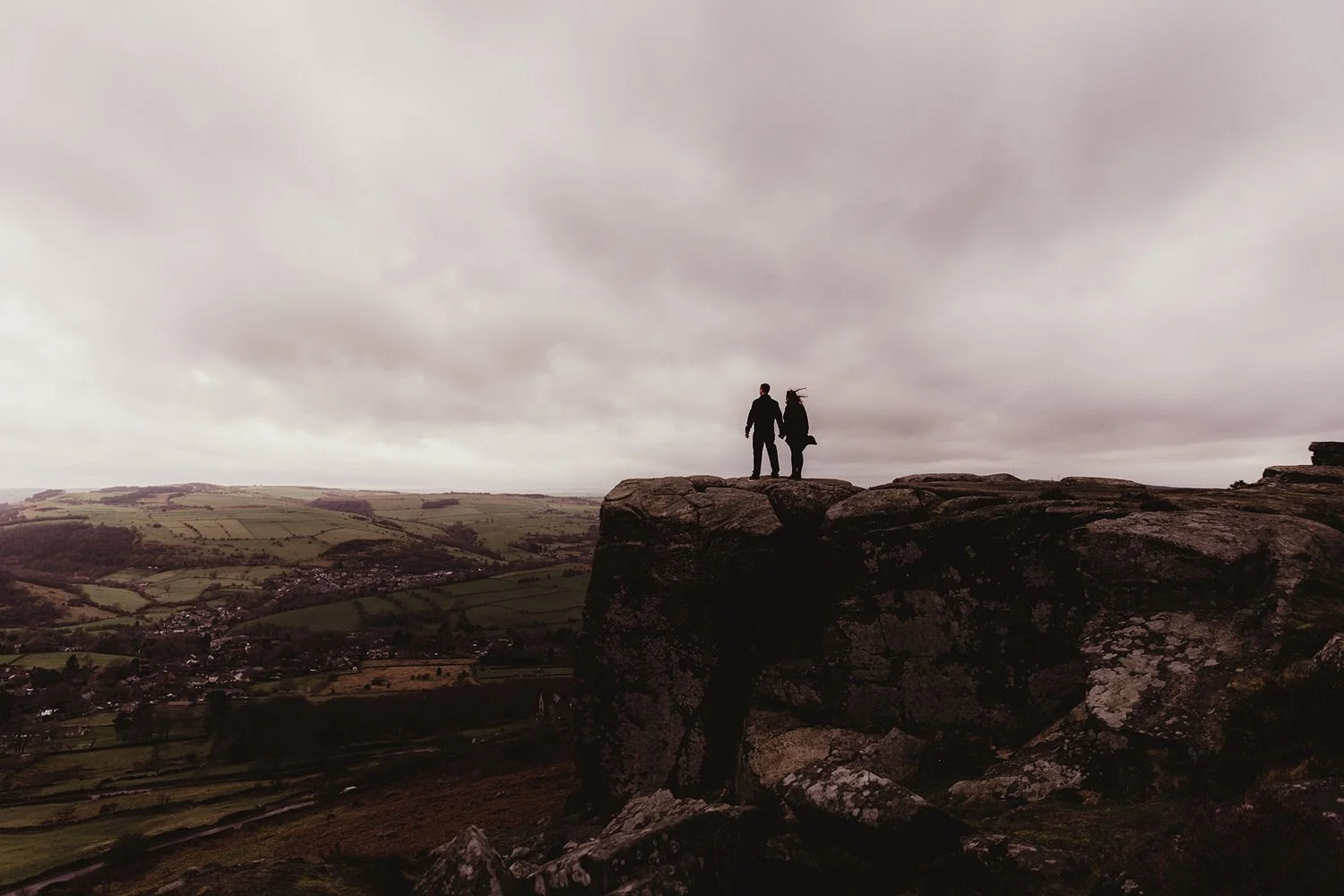 Silhouettes of a man and woman holding hands on a rocky cliff edge overlooking a rural valley with patchwork fields and small towns, under a cloudy sky.