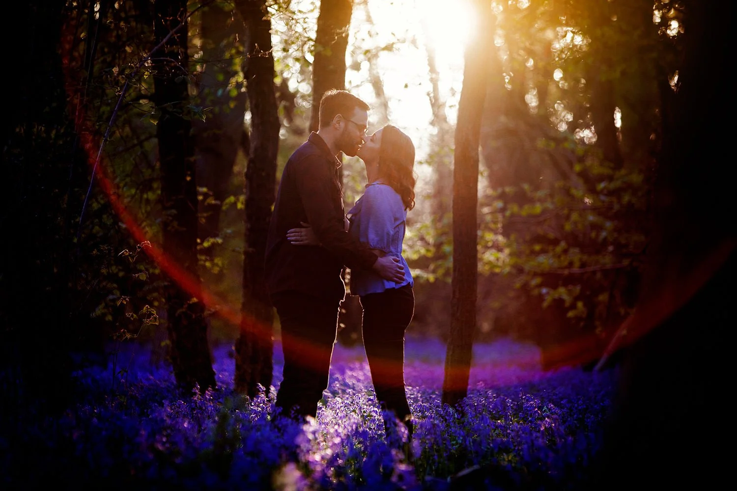 A couple standing close together in a forest during sunset, sharing a kiss. The ground is covered with bluebell flowers, and sunlight filters through the trees, creating a warm glow.