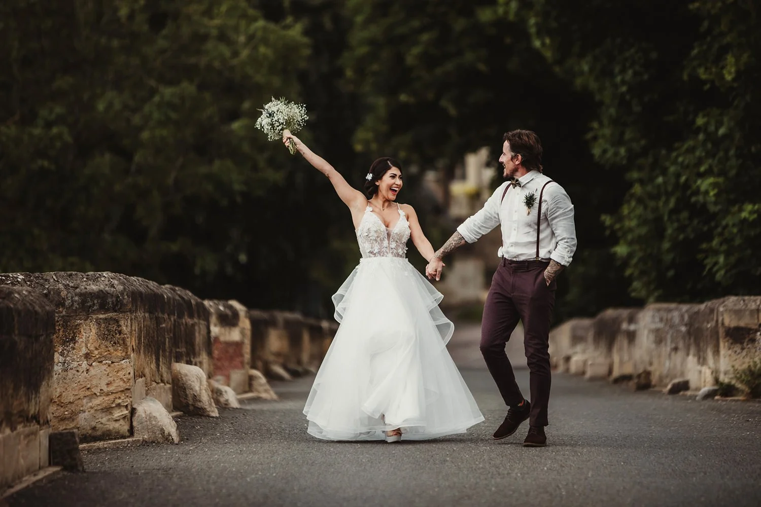 A bride and groom holding hands and dancing on a bridge, with the bride holding a bouquet, smiling, and the groom looking at her, surrounded by greenery.