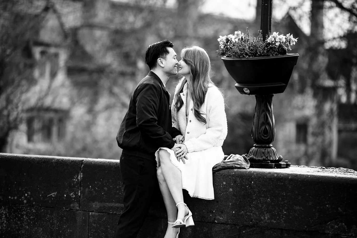 A black and white photo of a man and woman close together, smiling, holding hands, outside on a bridge or ledge, with trees in the background.
