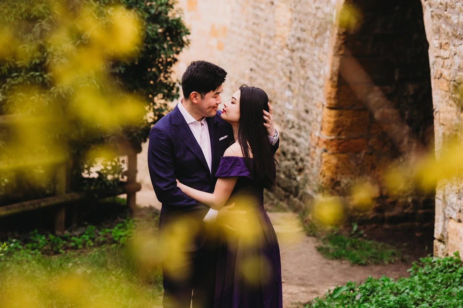 A young couple stands close together, gazing into each other's eyes, with the man gently holding the woman's face. They are dressed in formal attire, outdoors next to a stone wall, with blurred yellow flowers in the foreground.
