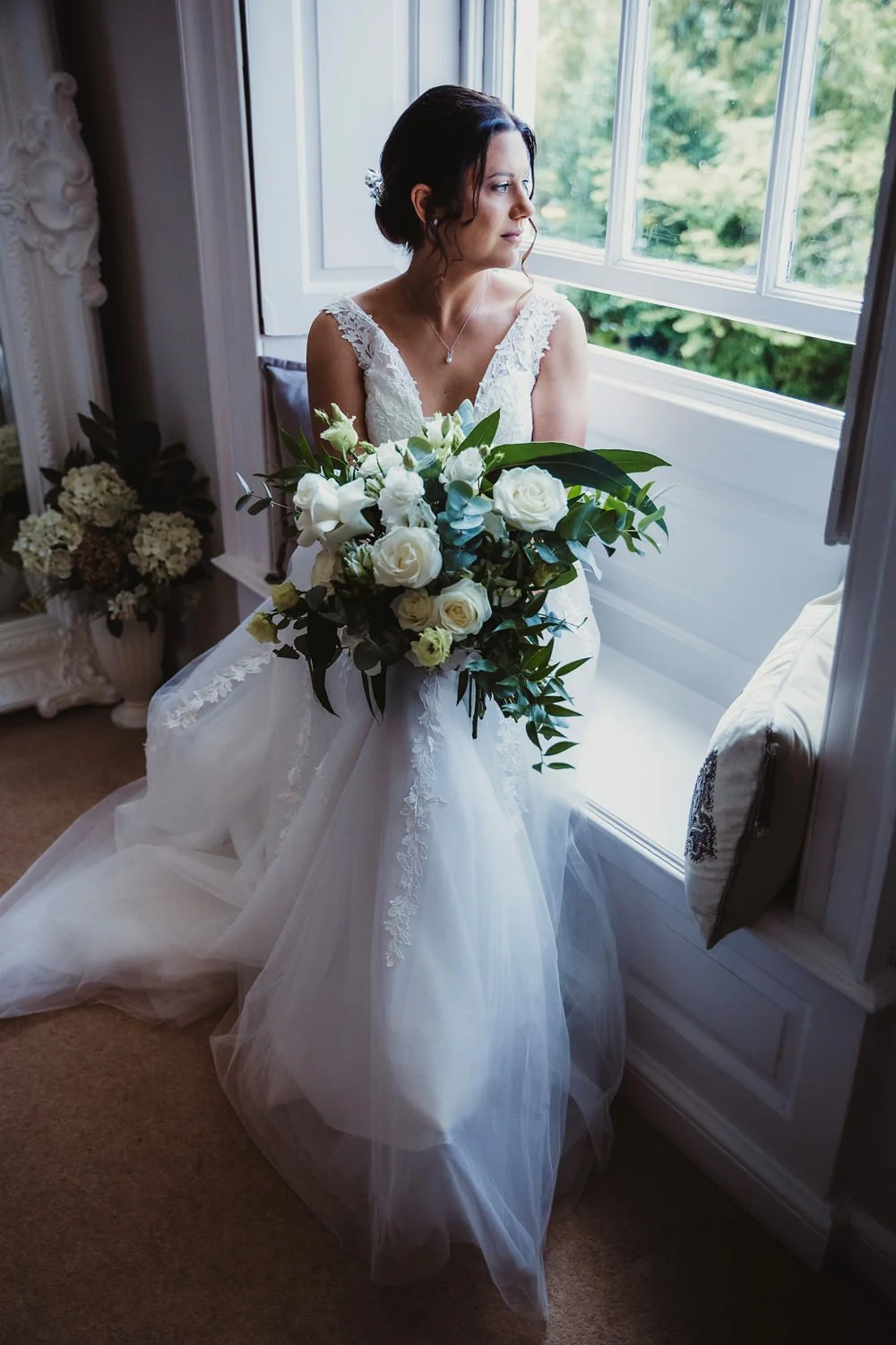 A bride in a white wedding dress holding a large bouquet of white roses and greenery, sitting near a window, gazing outside with a contemplative expression.