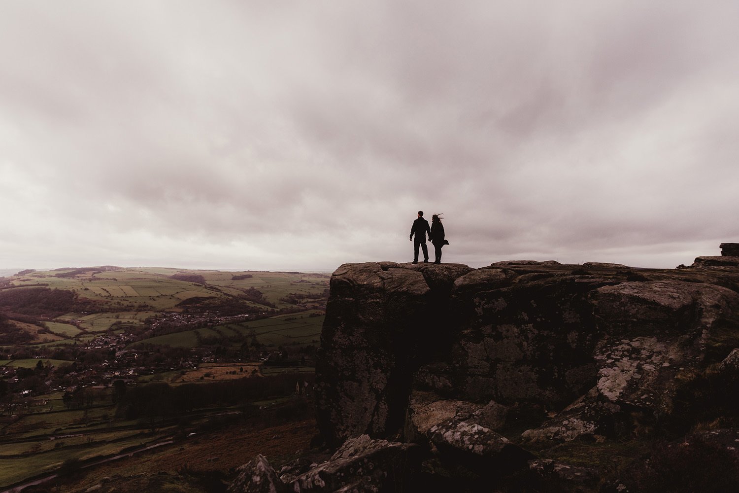 Silhouettes of two people stand on large rocks overlooking a valley with fields, trees, and a small town under a cloudy sky.