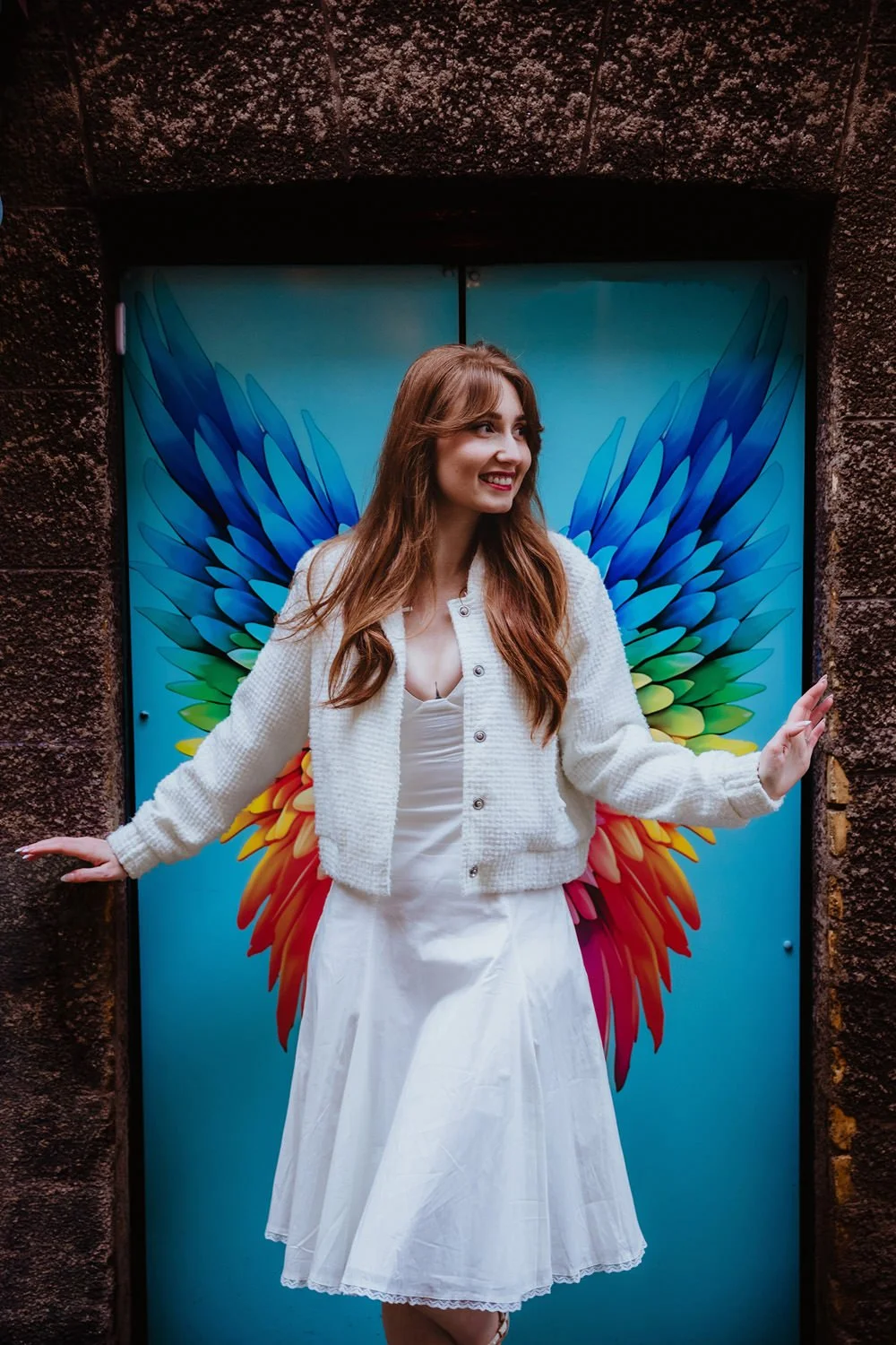A smiling woman with long brown hair wearing a white dress and a white cardigan stands in front of a colorful angel wings mural, giving the appearance of having wings.