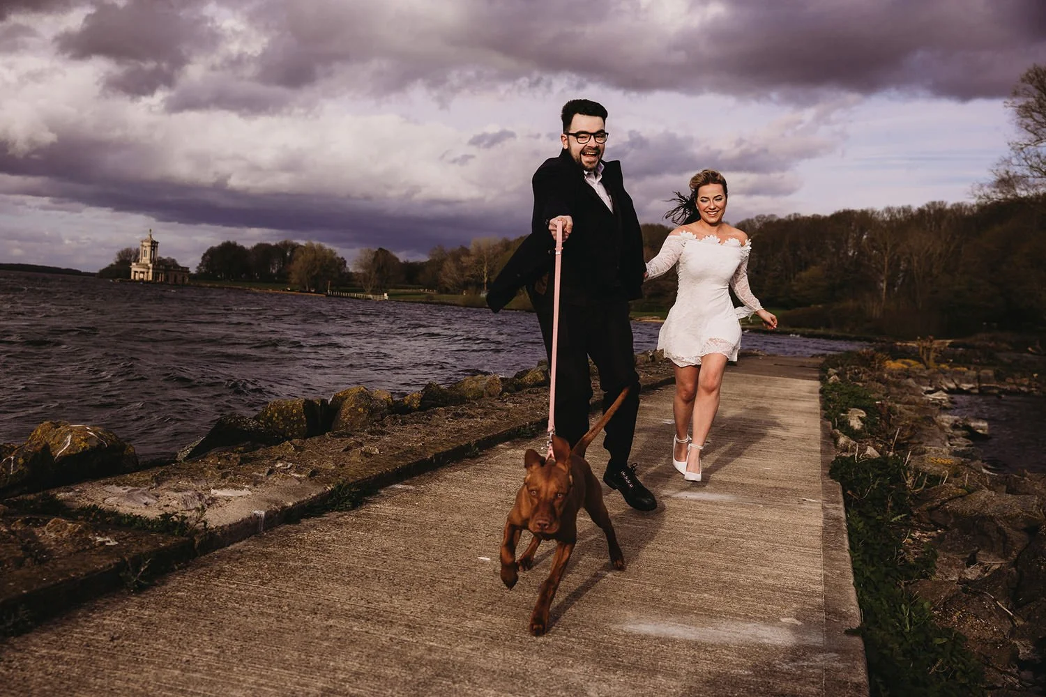 Engaged couple walking along a pathway beside a river while holding hands and running a brown dog on a leash. The weather is cloudy with dark clouds in the sky.